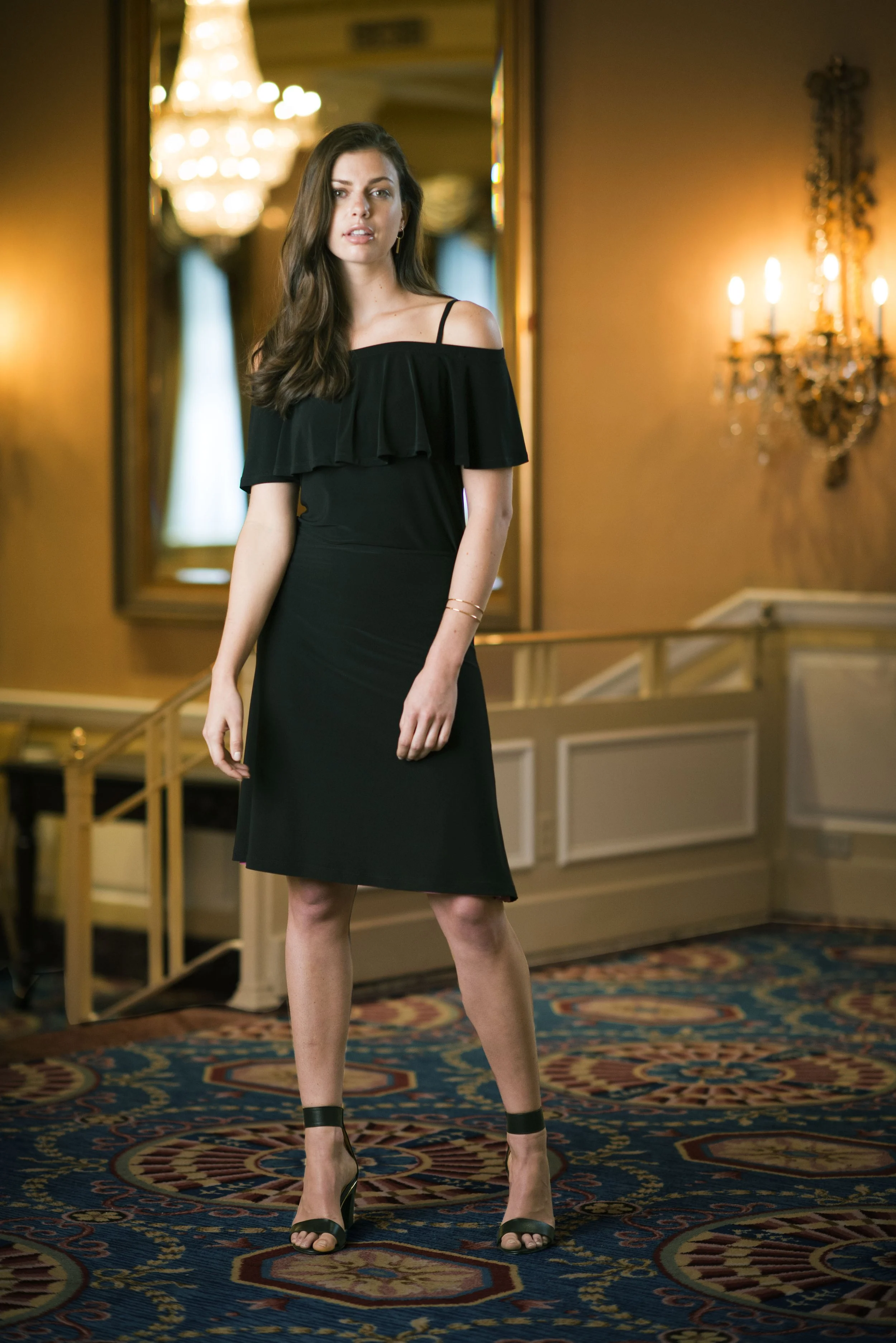 A young woman in a black off-the-shoulder dress and high heels stands in an elegant, warmly lit room with a chandelier, patterned carpet, and ornate wall sconces.