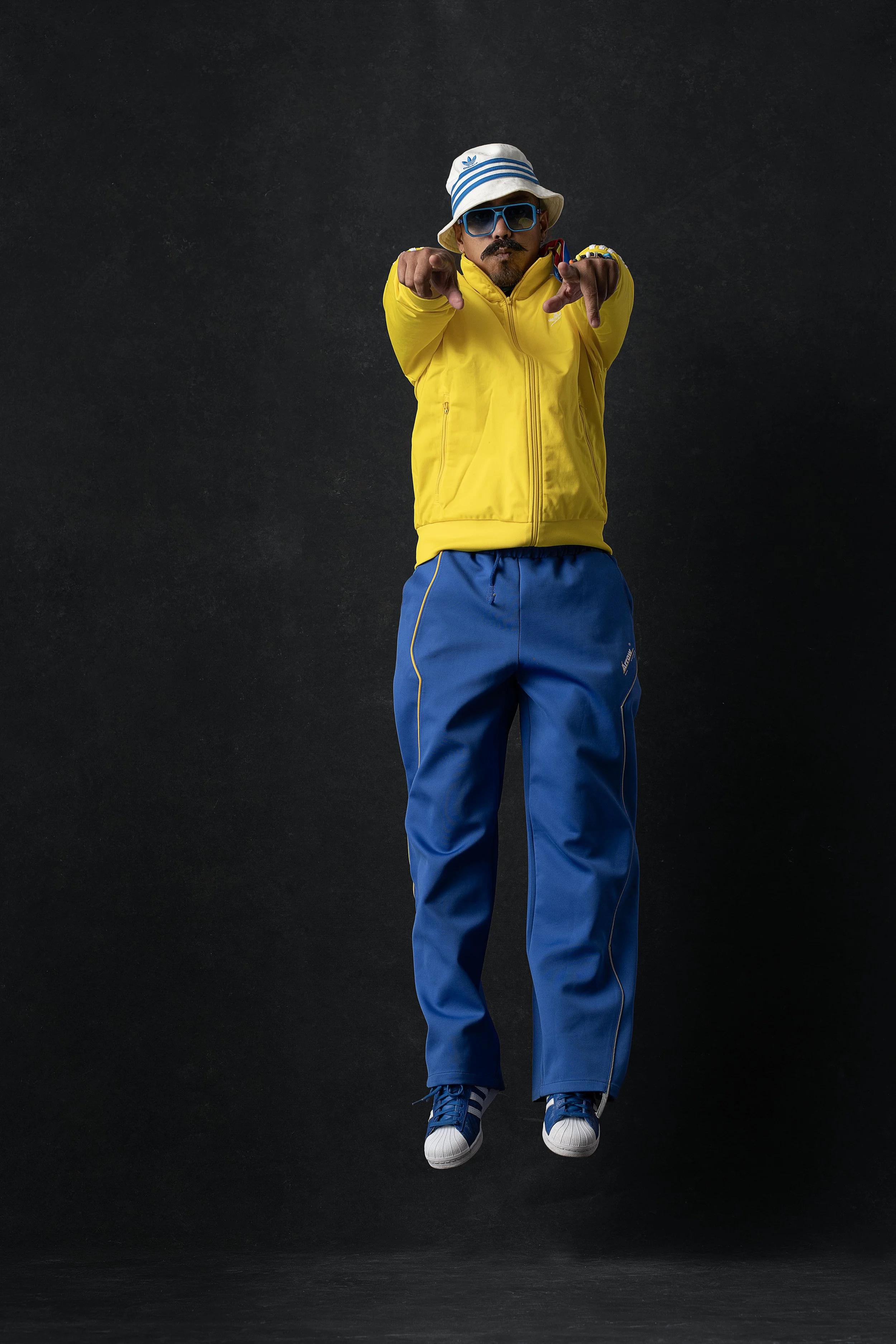 A man wearing a yellow jacket, blue pants, white sneakers, a bucket hat, and sunglasses is jumping in the air against a dark background, pointing upfront.