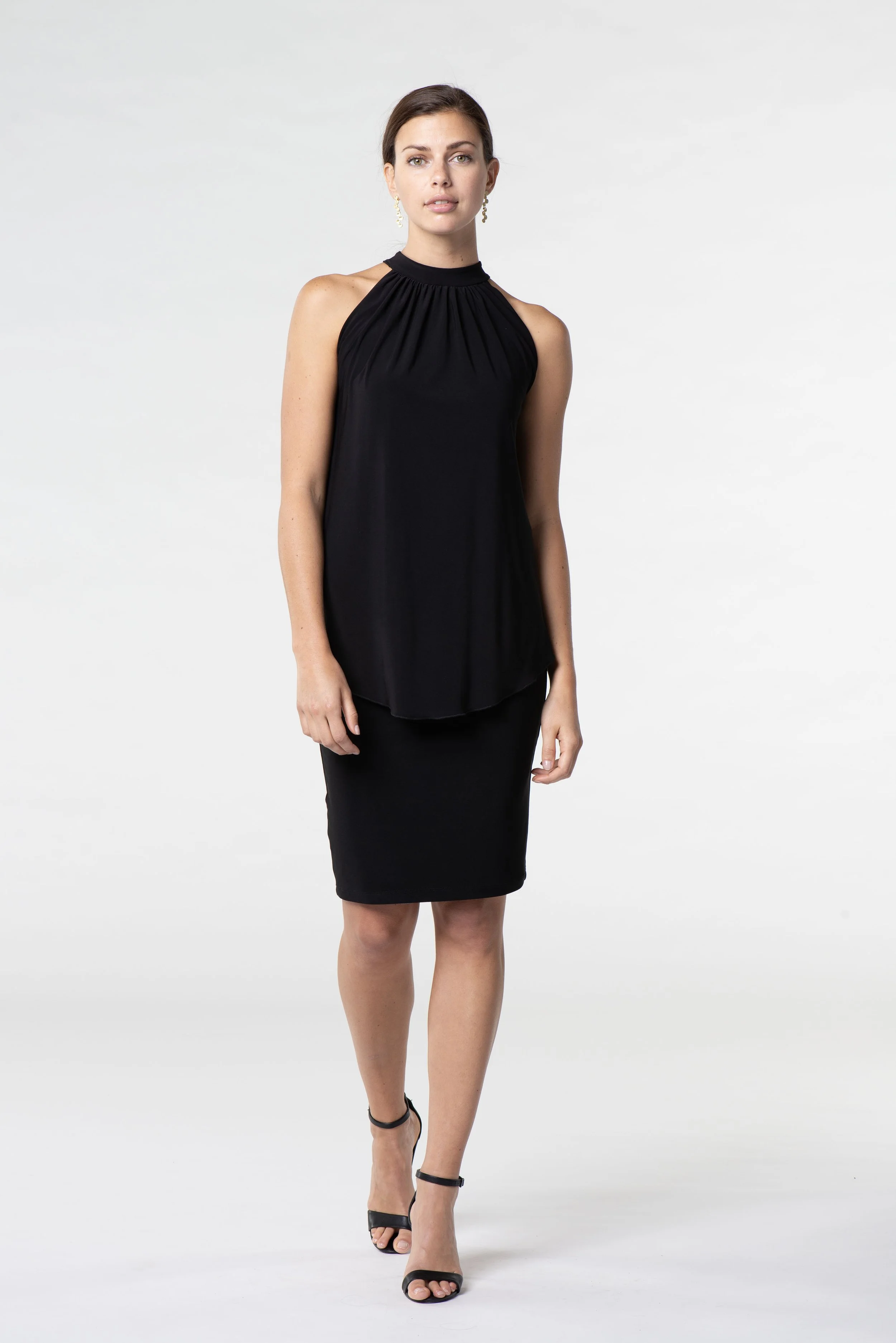 A woman wearing a black sleeveless top with gathered neckline, black skirt, and black high heels, standing against a white background.