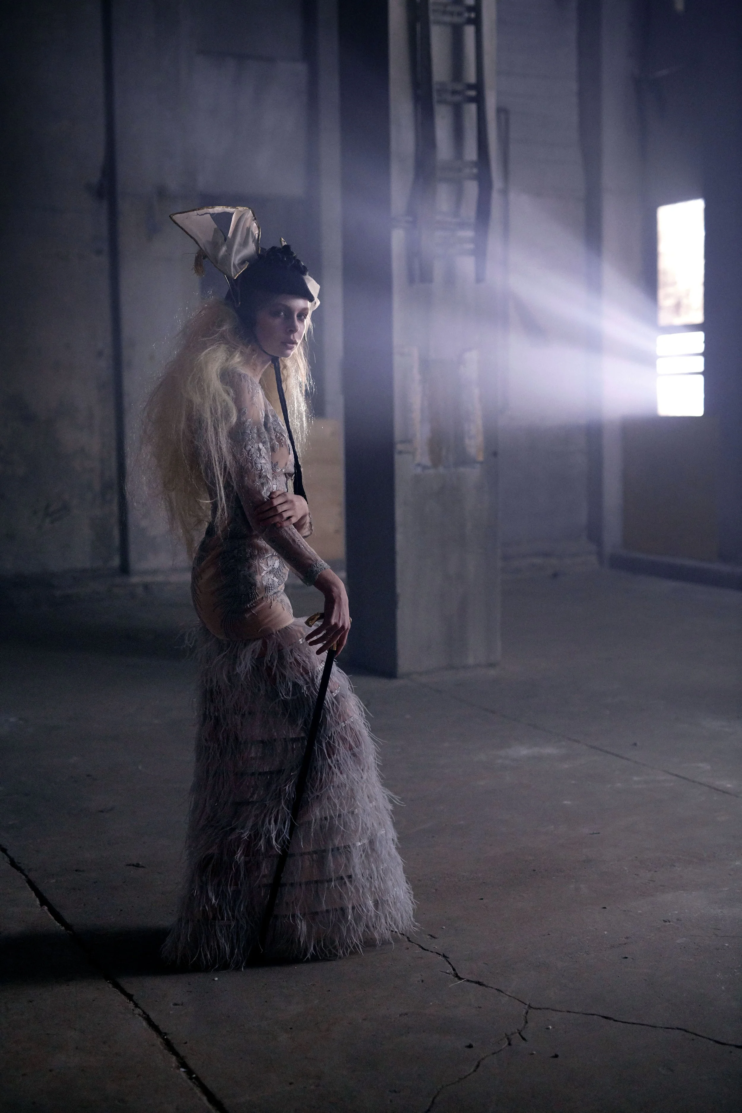 A woman dressed in an elaborate costume with a feathered headpiece, standing in a dimly lit, industrial-style space with beams of light streaming through windows.