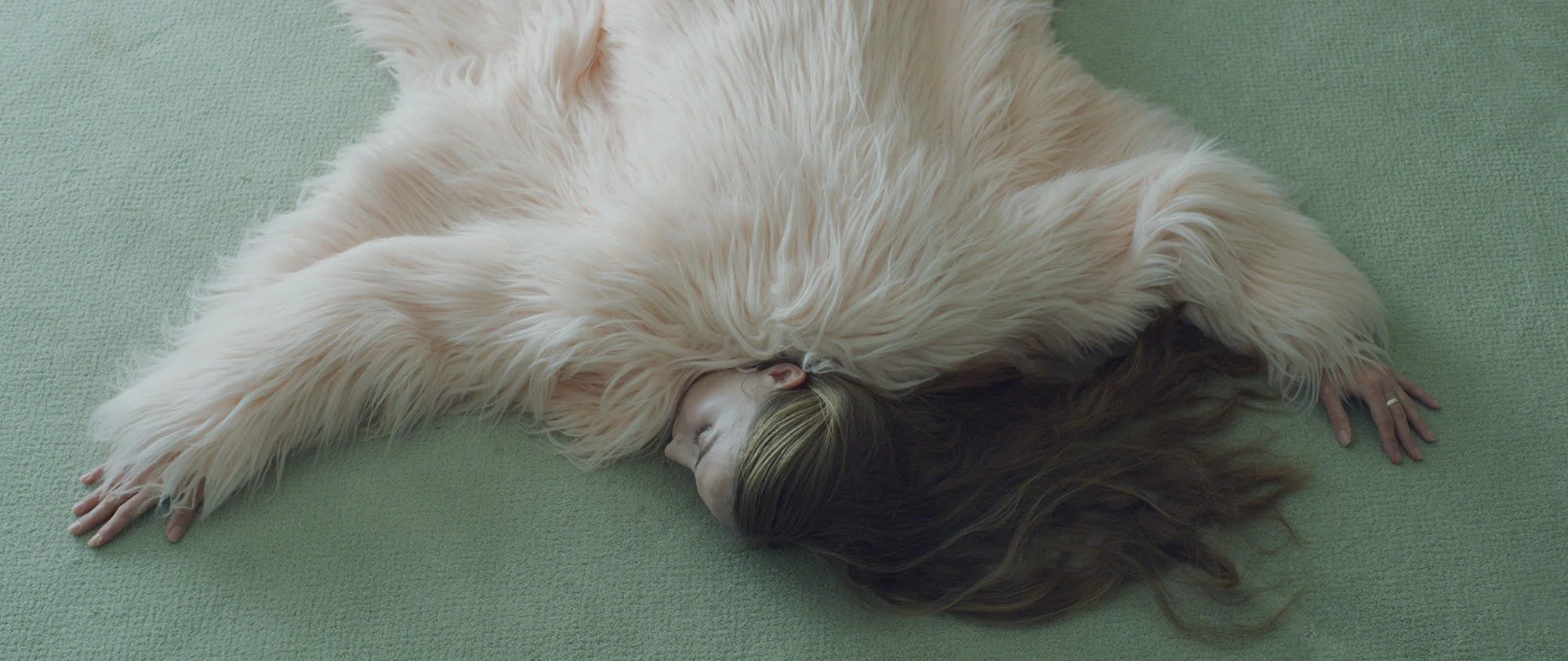A woman lying on her side on a green mat with her arms outstretched, covered in a furry, cream-colored blanket.