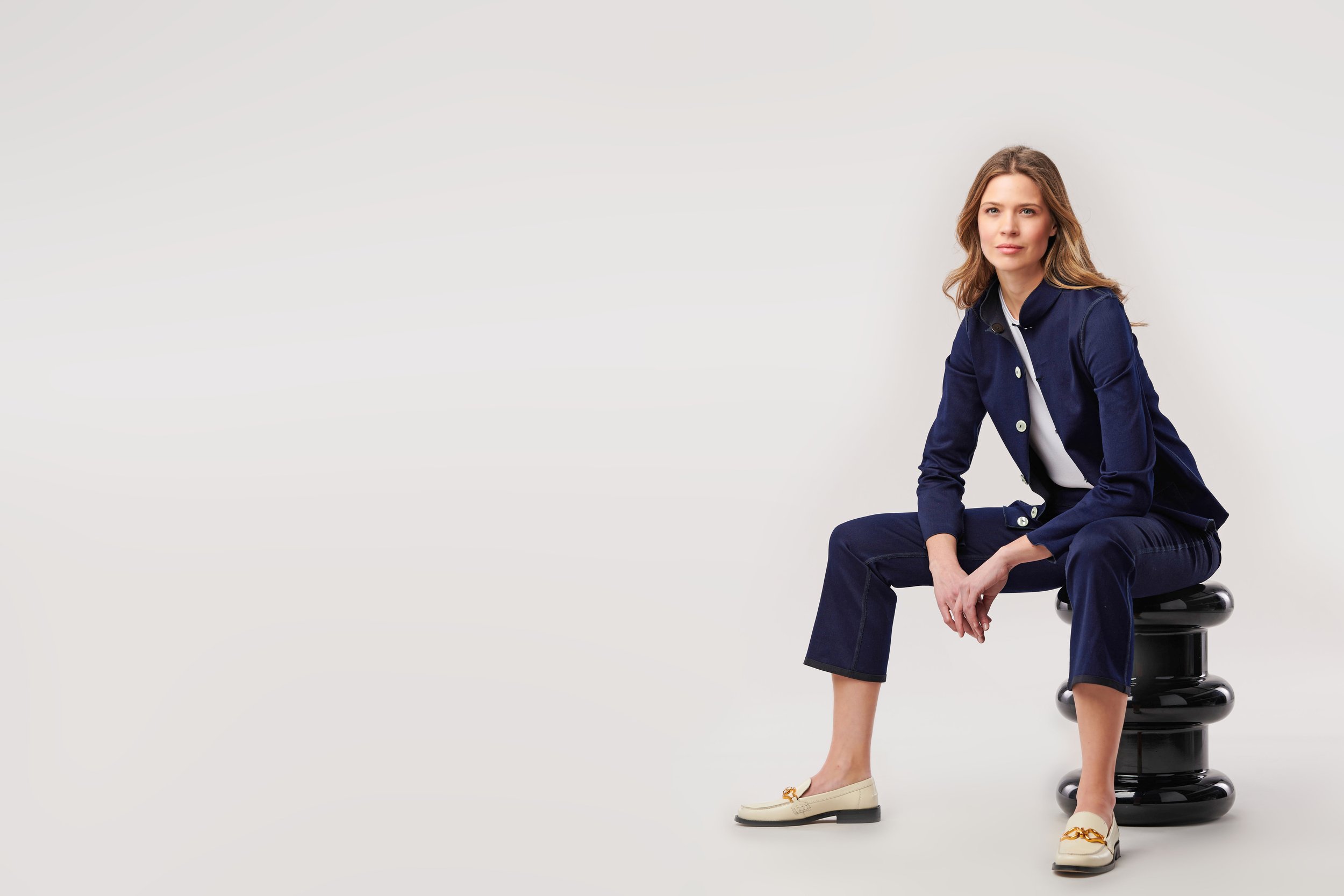 A woman with wavy brown hair sitting on a black stacked stool against a plain white background. She is wearing a navy blue suit and beige loafers with a gold detail, and poses with one foot on the ground and one leg crossed over, looking slightly to the right.