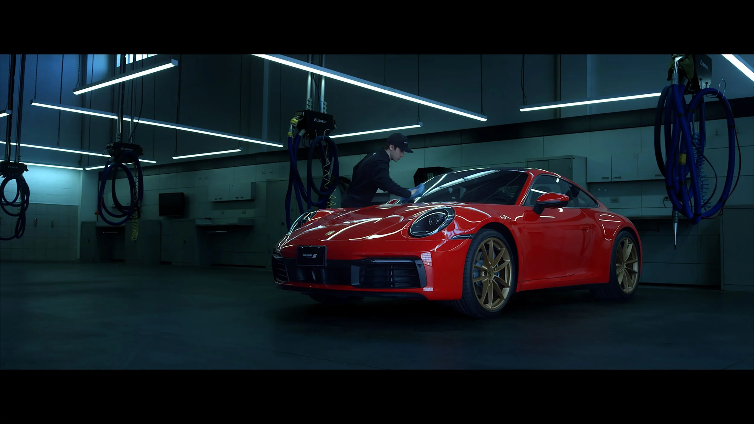 A person in a black uniform and cap cleaning a red sports car with gold wheels inside a modern, well-lit garage with equipment hanging on the walls.
