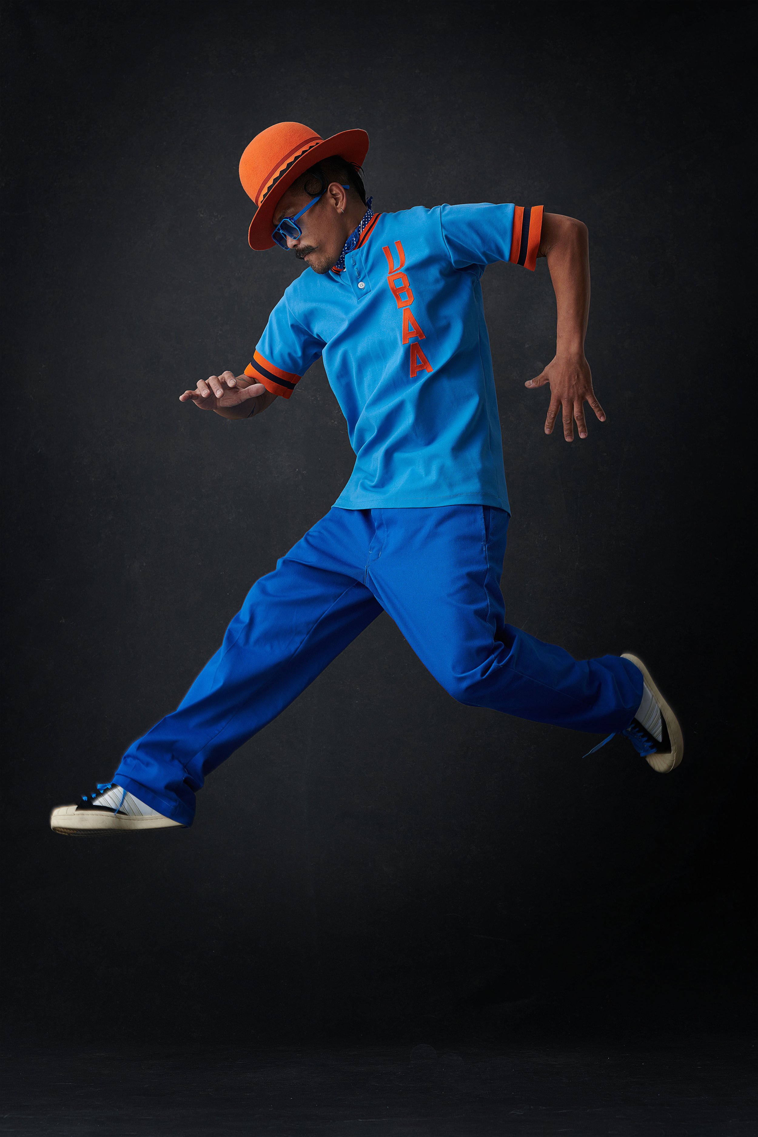 A man wearing a blue and orange baseball-style outfit, with a large orange hat, blue glasses, and white sneakers, captured mid-motion against a dark background.