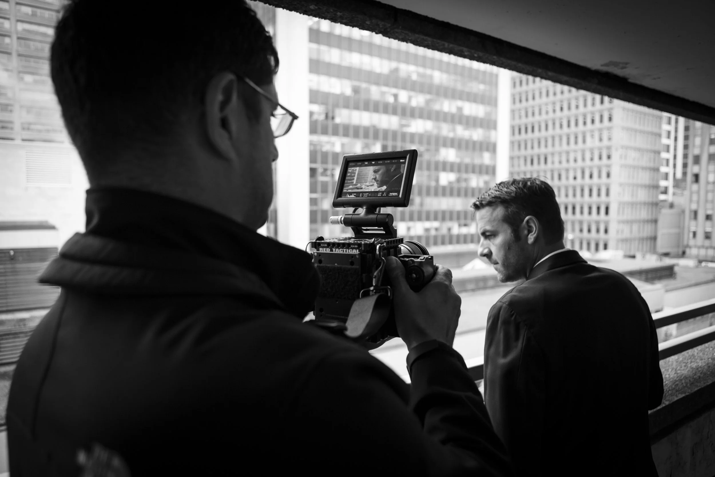 A man operating a camera on a city balcony, with another man leaning over the balcony railing, high-rise buildings in the background.