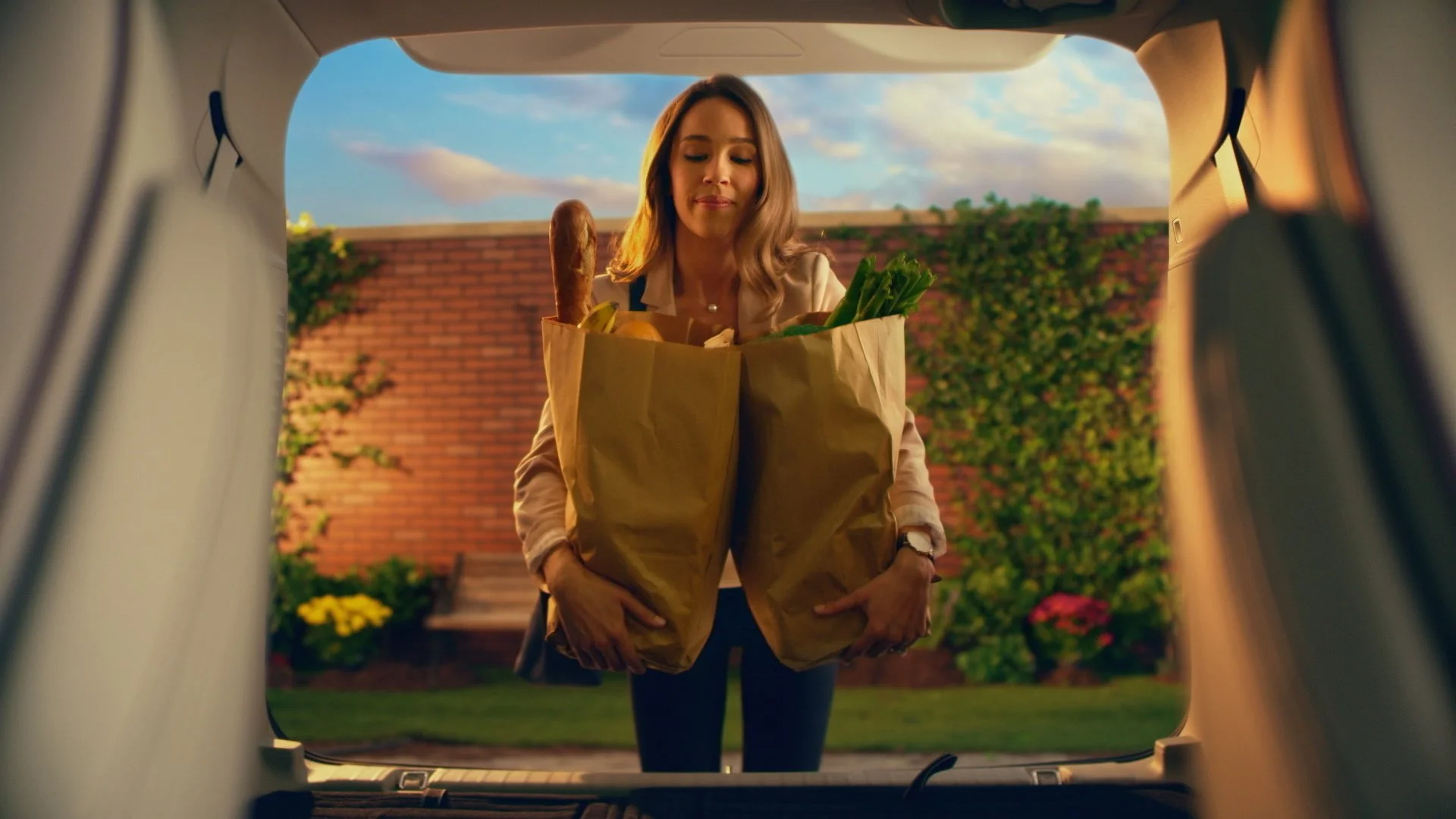 Woman unloading grocery bags into the trunk of a car in a backyard with a brick wall and flowering plants.