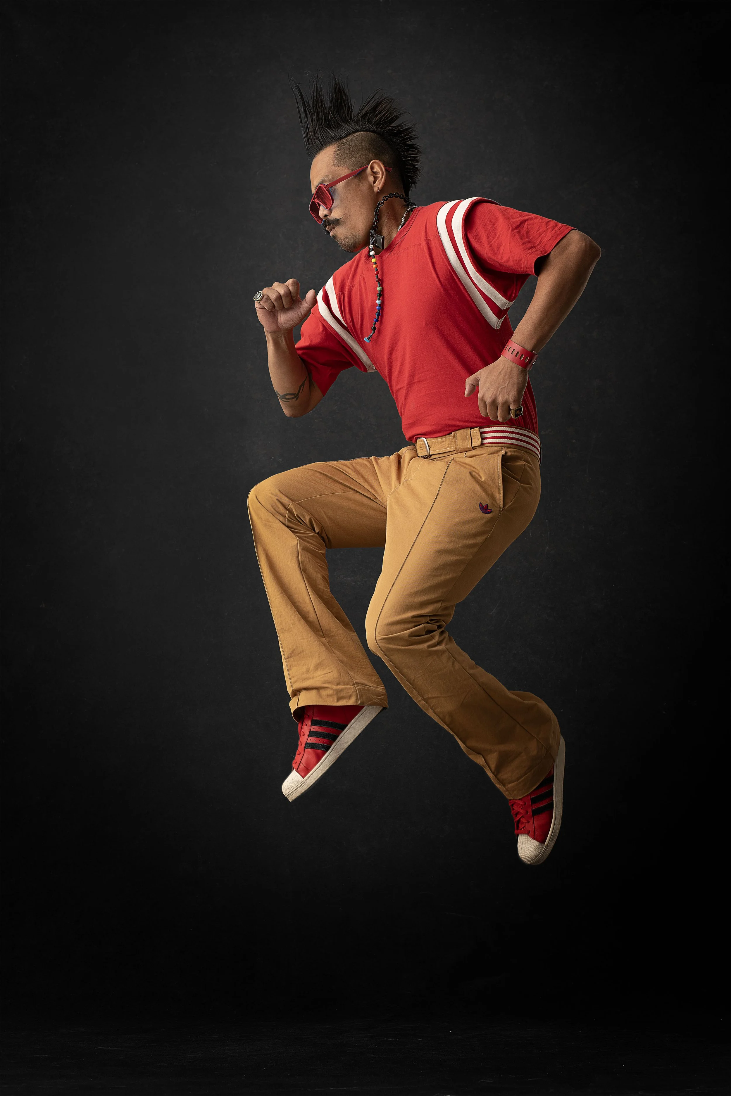A man with a mohawk hairstyle, wearing red sunglasses, a red sports jersey, khaki pants, and red sneakers, appears to be jumping or dancing against a black background.