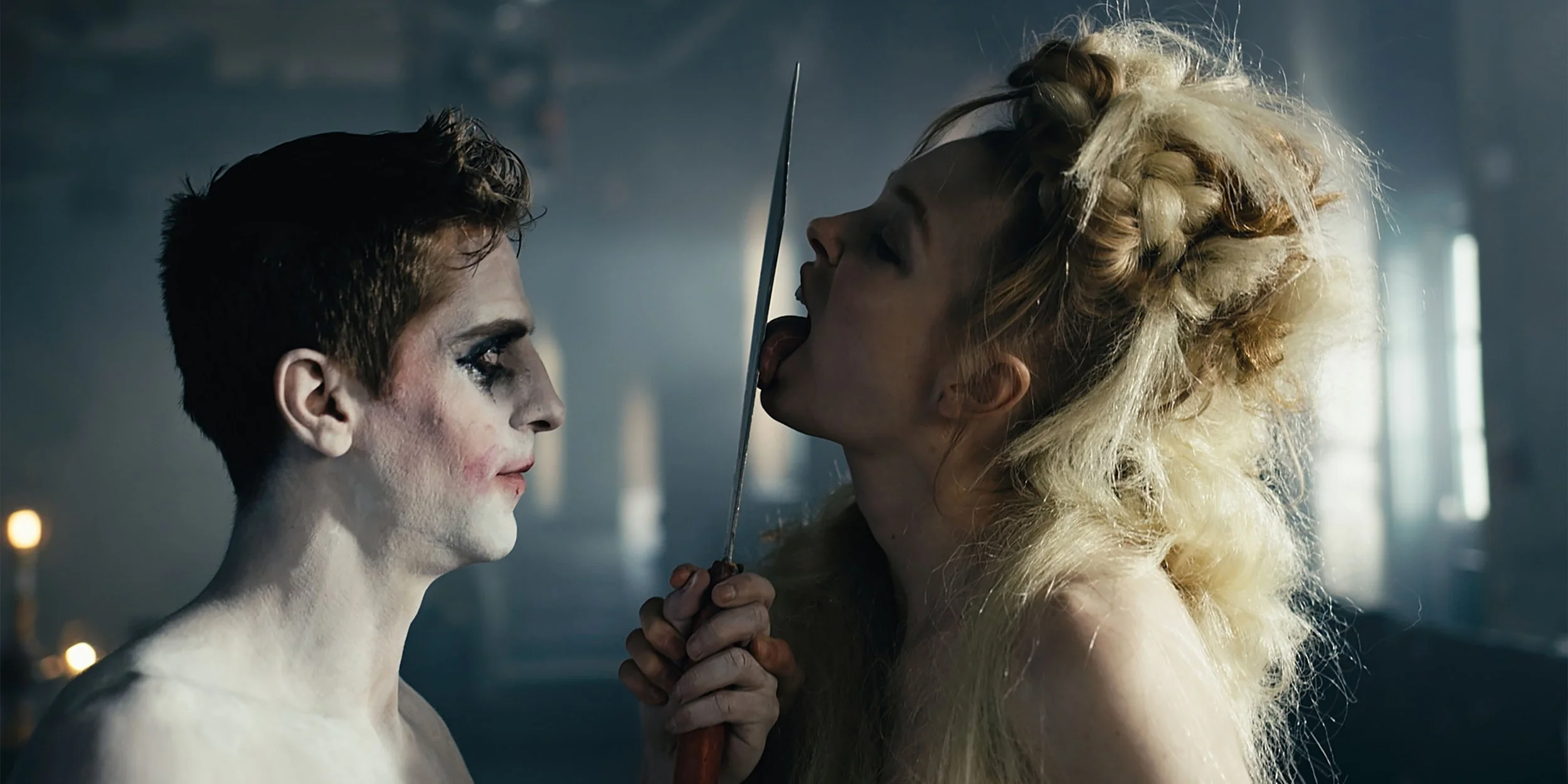 Two women with intense makeup and wild hairstyles stand close, holding a sword vertically between them in a dimly lit room with a moody atmosphere.