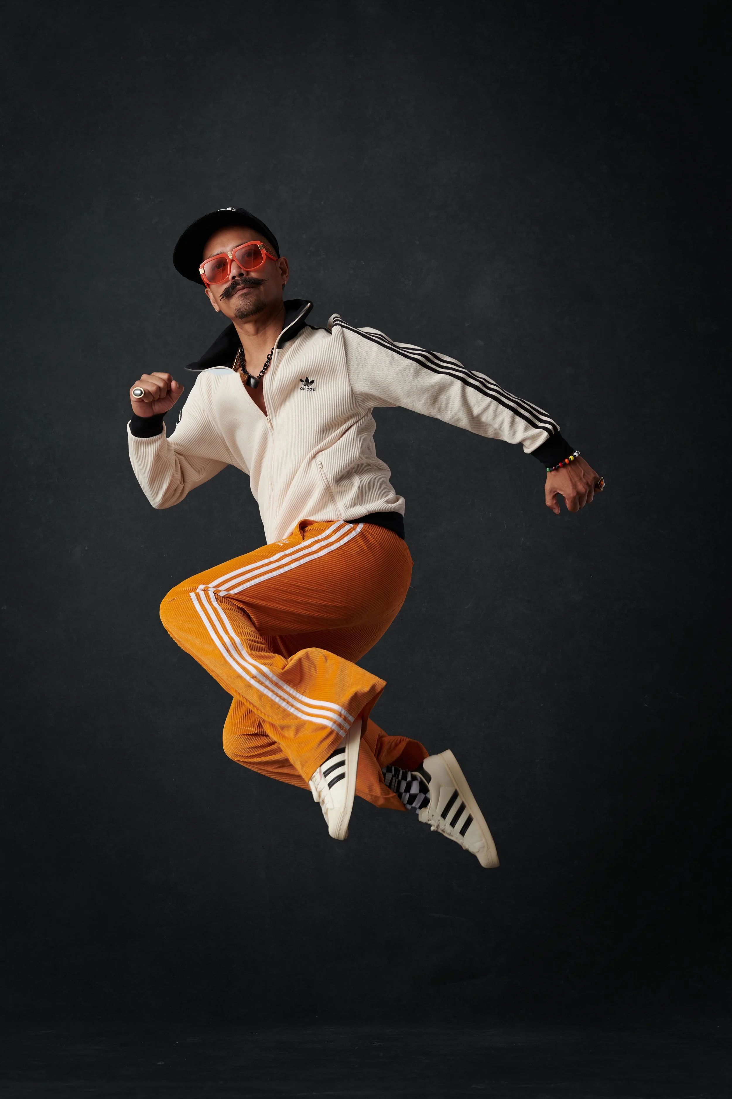 A man with a mustache and beard is mid-air with a black background behind him. He is wearing a beige Adidas zip-up jacket with black stripes, orange pants with white stripes, white Adidas sneakers, a black cap, orange tinted glasses, and various accessories including rings, necklaces, and bracelets.