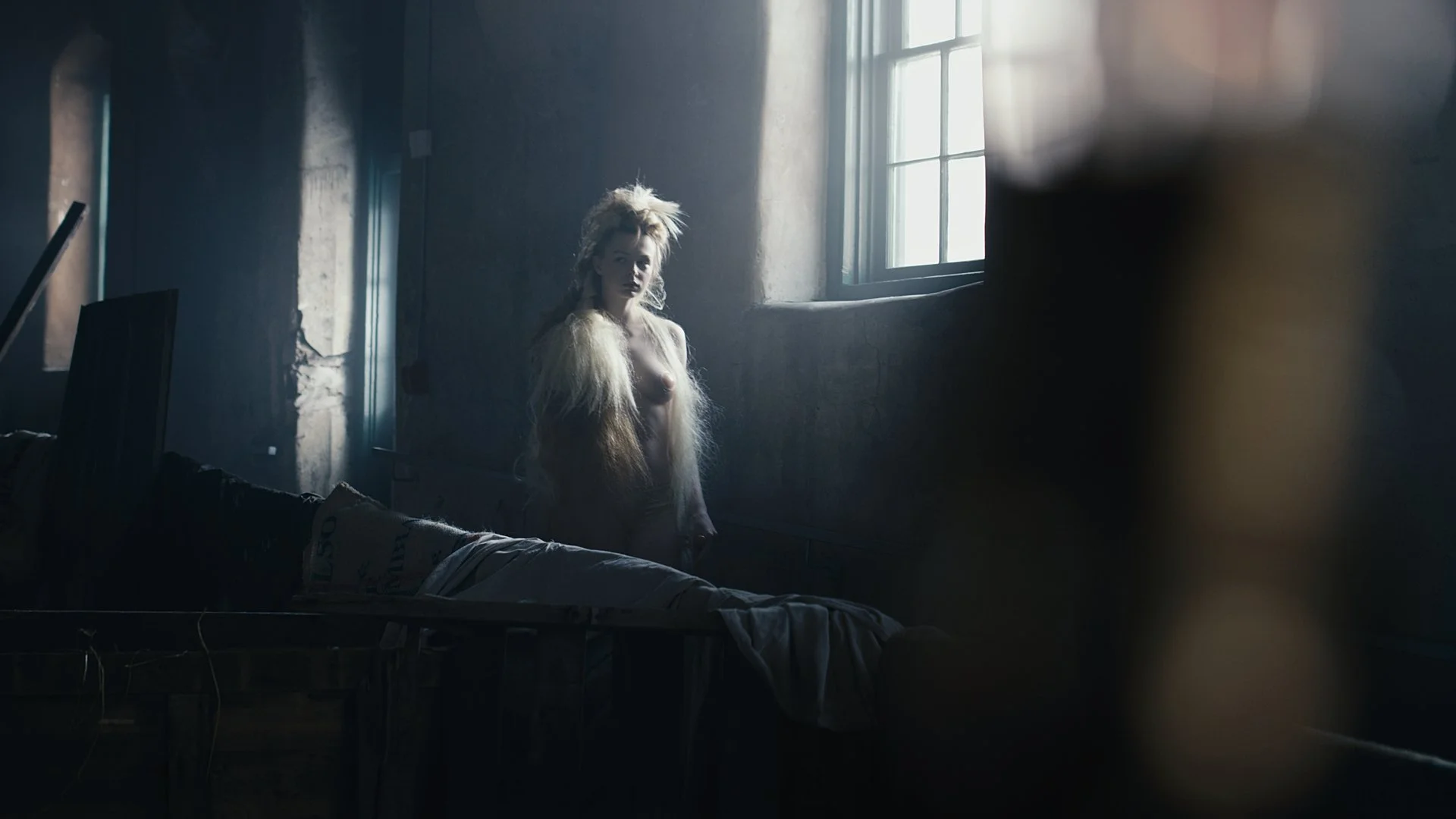A woman with long, wild hair standing partially undressed in a dimly lit, rustic room with a large window letting in natural light.