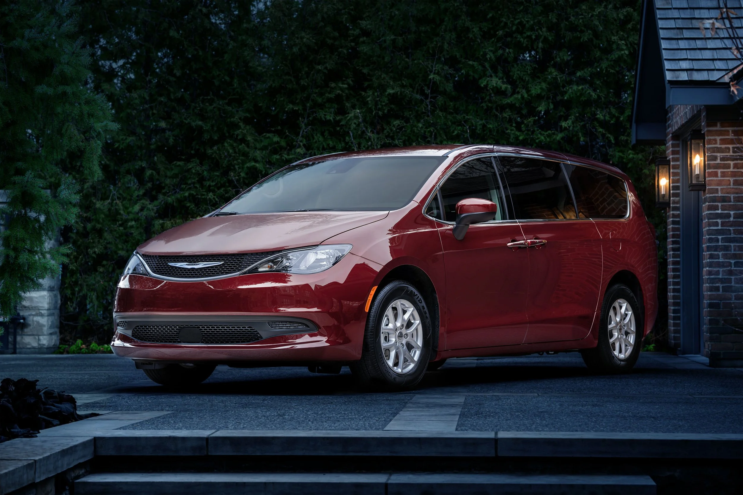 A red minivan parked in front of a brick house with lantern-style lights, surrounded by green trees during dusk.