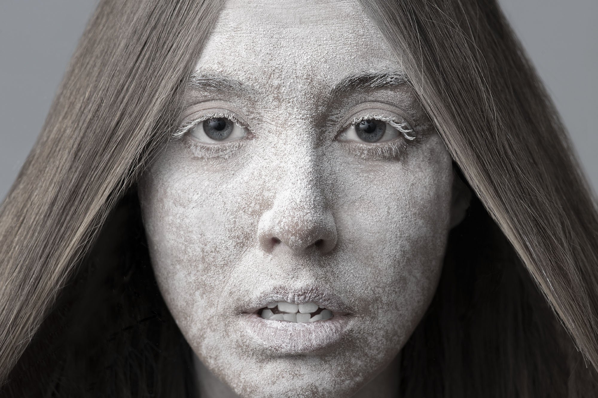 A close-up of a woman's face with silver paint on her skin, light brown hair, and blue eyes, with a neutral background.