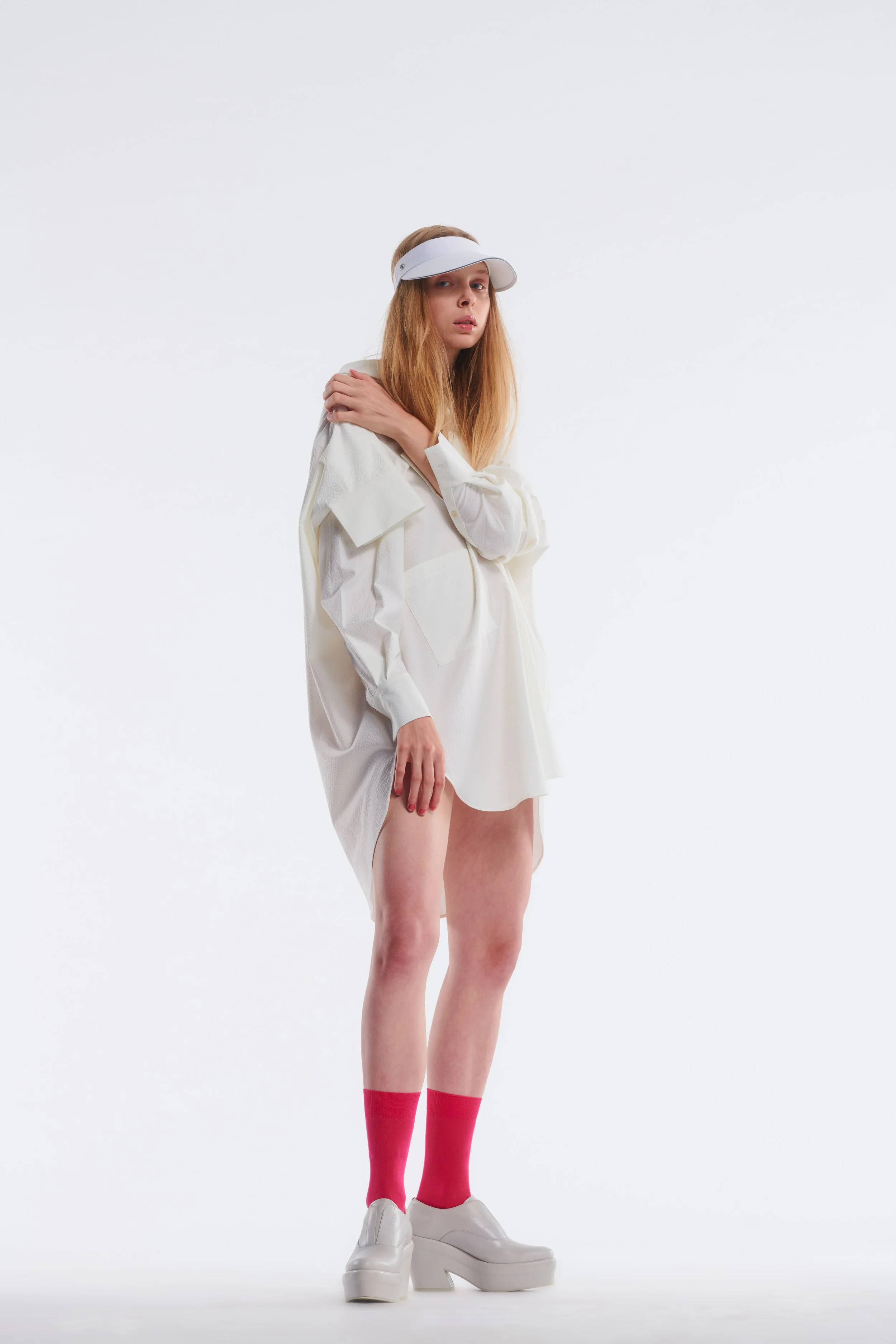 A young woman with long blonde hair standing against a plain white background. She is wearing a white visor, a light-colored oversized shirt, pink knee-high socks, and white platform shoes.