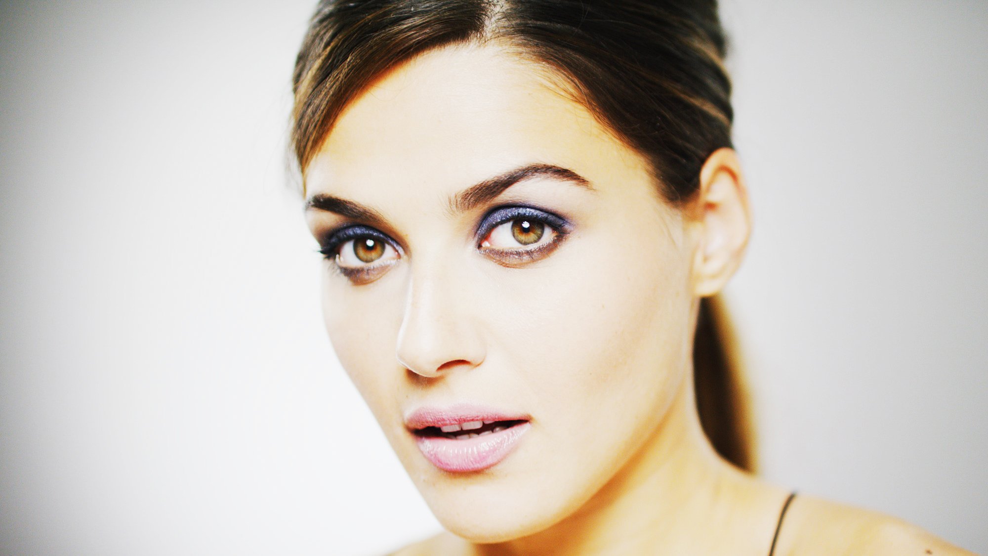 Close-up of a woman with brown hair and brown eyes, wearing eye makeup, looking slightly to the side, with a neutral expression.