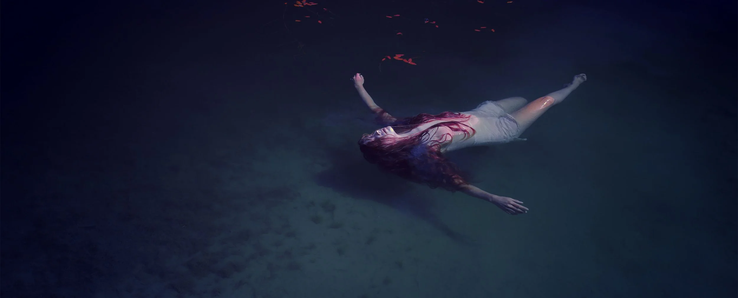 A woman with long red hair floats face-up in dark water, wearing a light-colored dress. The water is dark and the scene has a surreal, peaceful atmosphere.