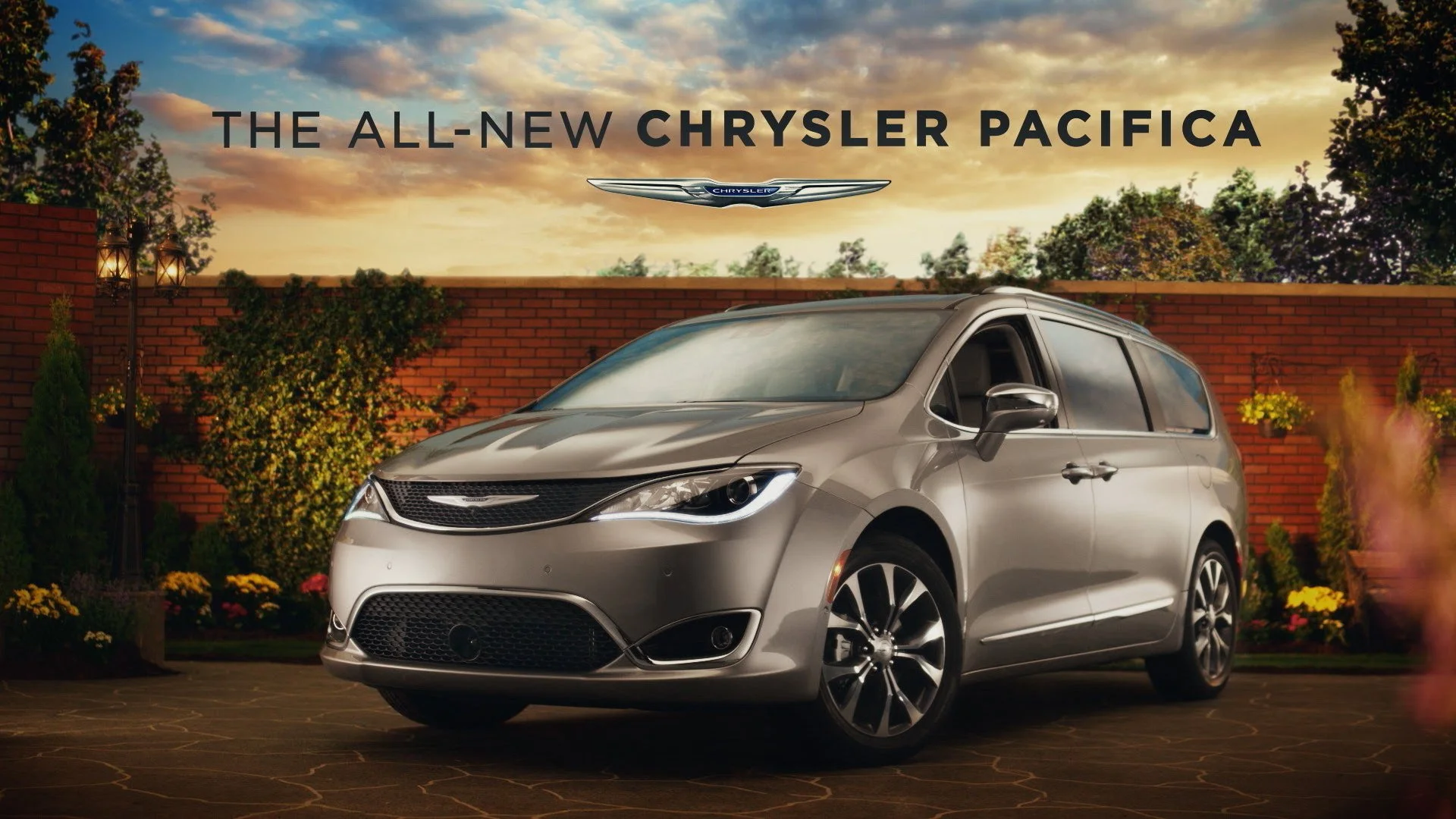 Silver Chrysler Pacifica minivan parked in a driveway during sunset with a brick wall, trees, and plants in the background, advertising the all-new Chrysler Pacifica.
