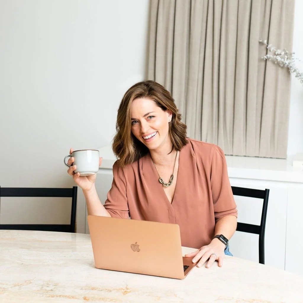 A woman sitting at a table with a laptop, smiling and holding a mug.