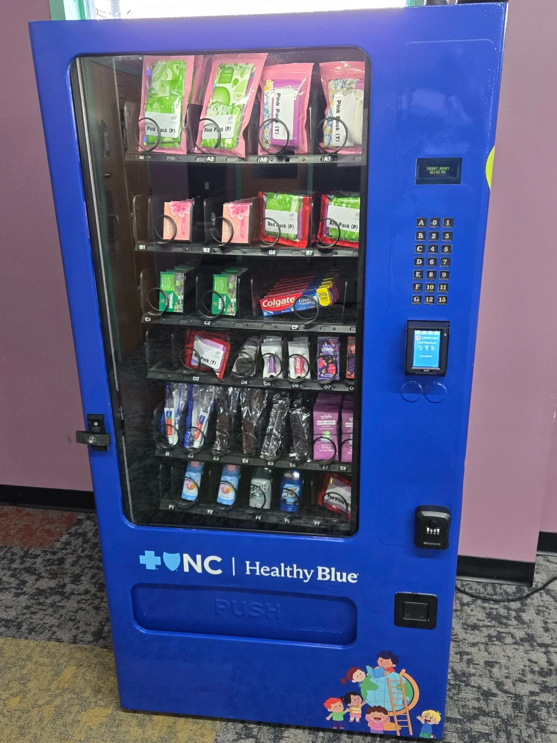 A blue vending machine filled with health and wellness products including packs of gum, mints, and toothpaste. The vending machine has a digital screen and a keypad for selection, located indoors on a carpeted floor.