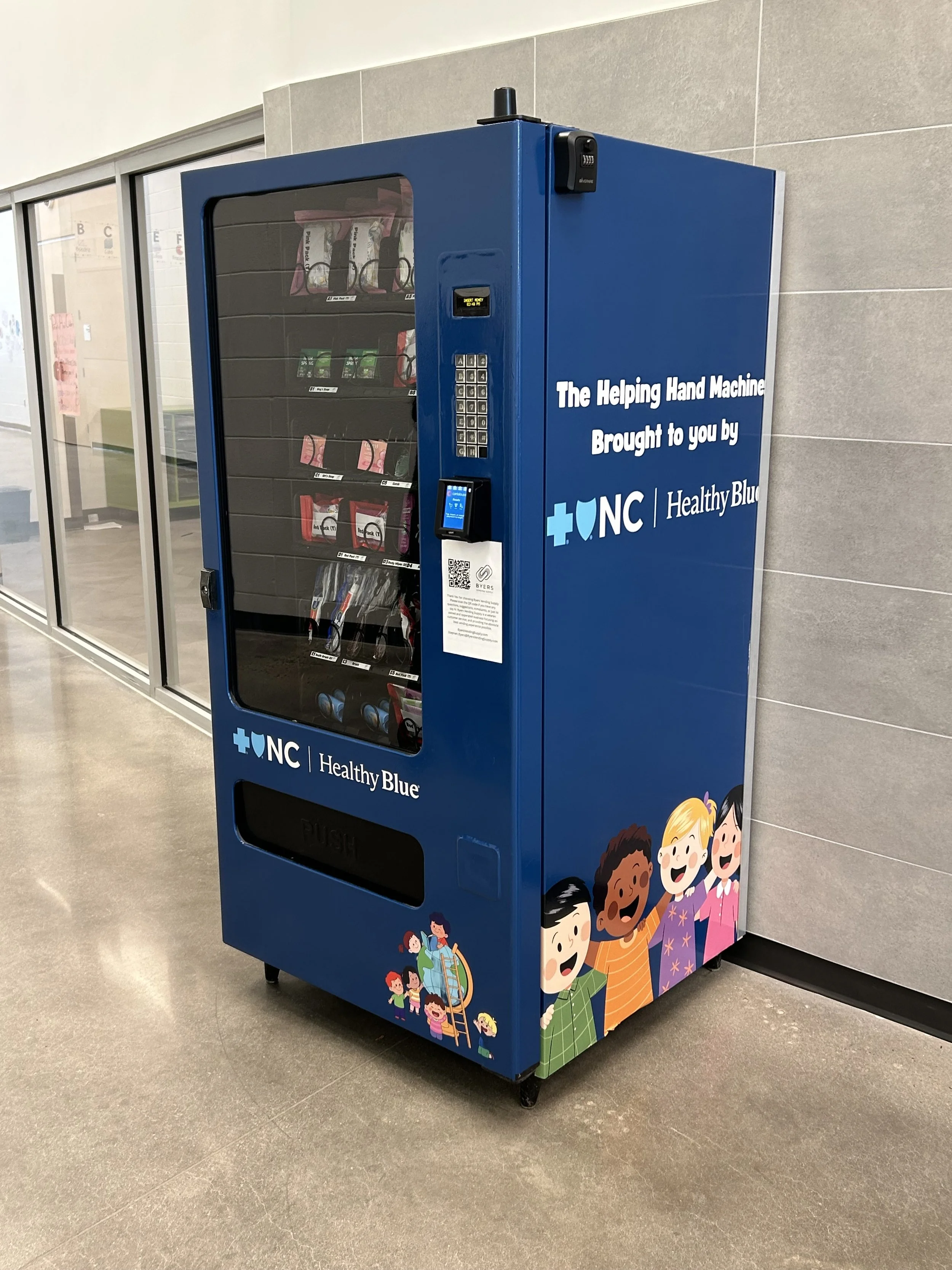 A vending machine named 'The Helping Hand Machine' with a blue exterior, located in a school or community center, decorated with cartoon children, and offering snacks and items in glass compartments.