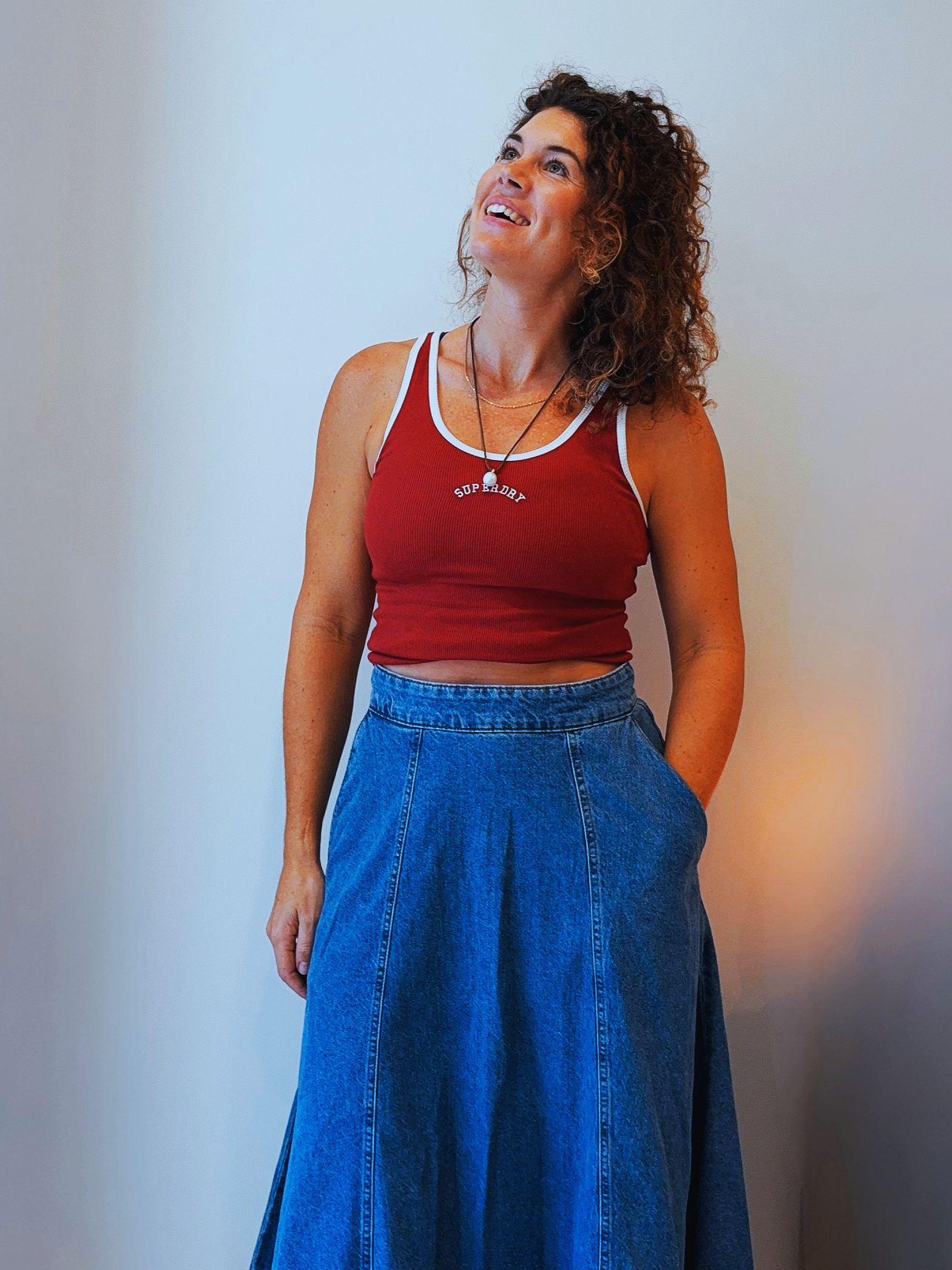 A woman with curly brown hair wearing a red tank top with white trim and a long denim skirt, standing against a plain wall and gazing upward.