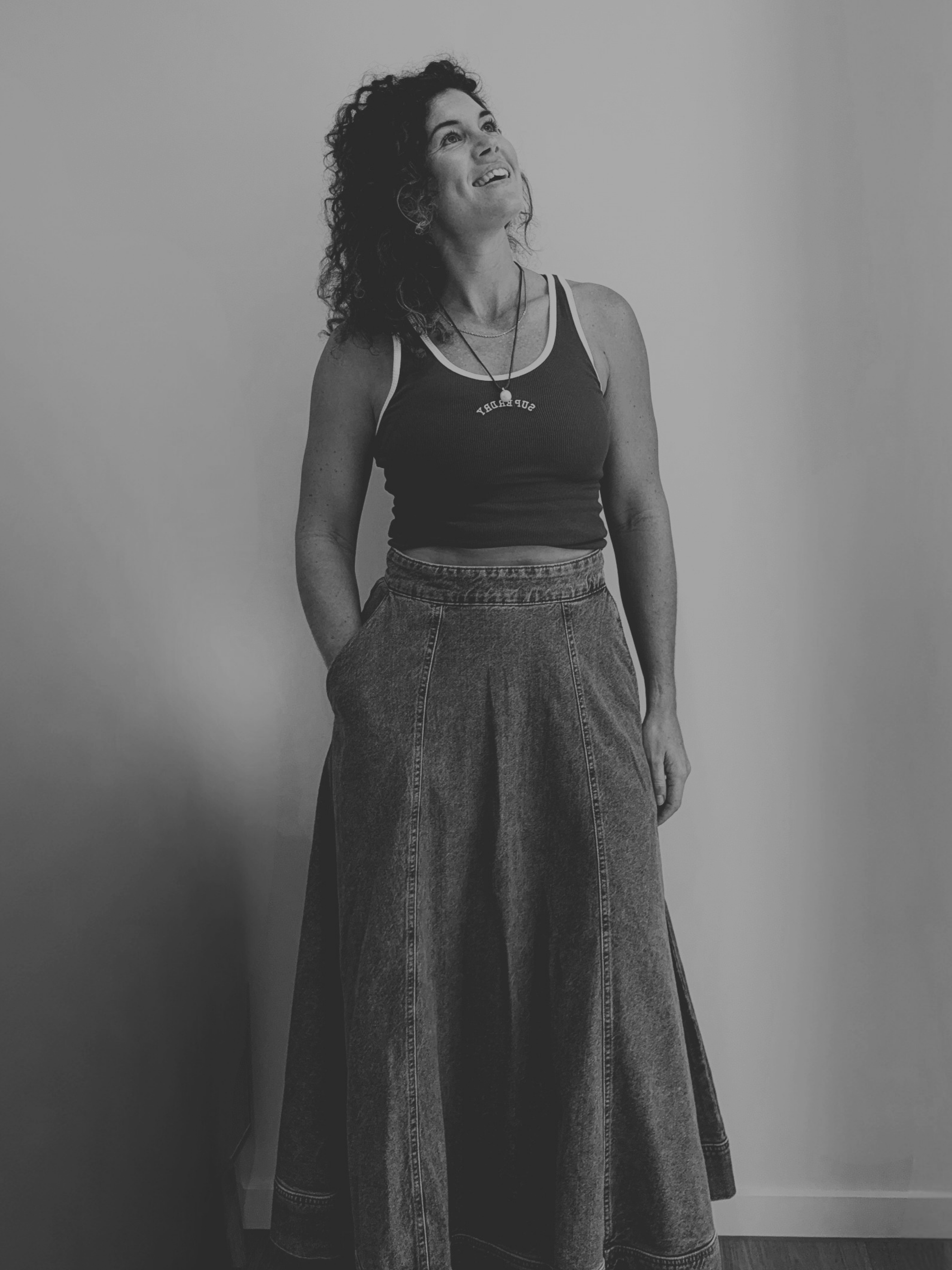 A woman with curly hair wearing a tank top and wide-leg jeans, standing against a plain wall, smiling and looking up.