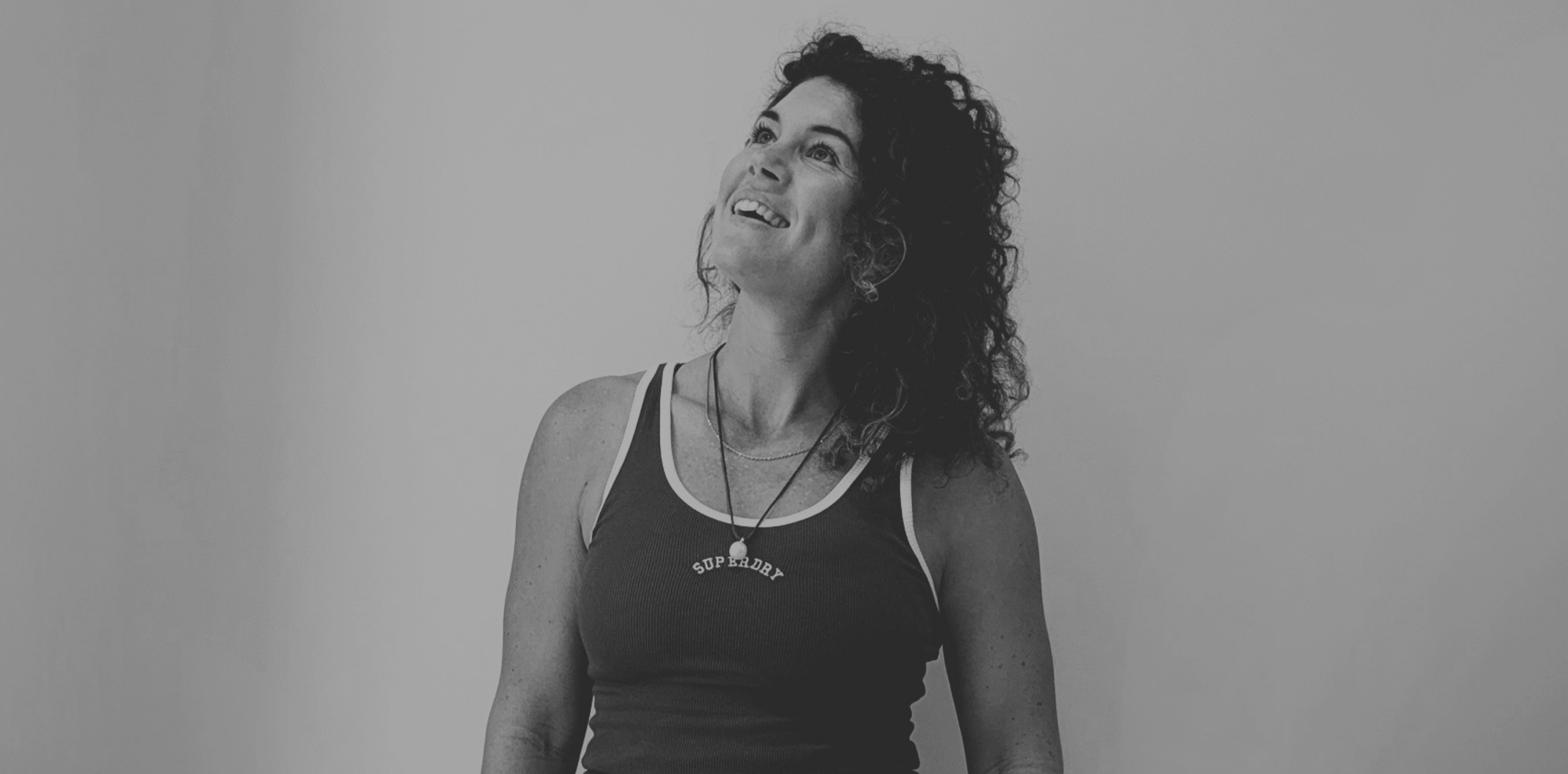 A woman with curly hair and freckles wearing a tank top with 'Superdry' written on it, looking upwards with a smile, standing against a plain background.