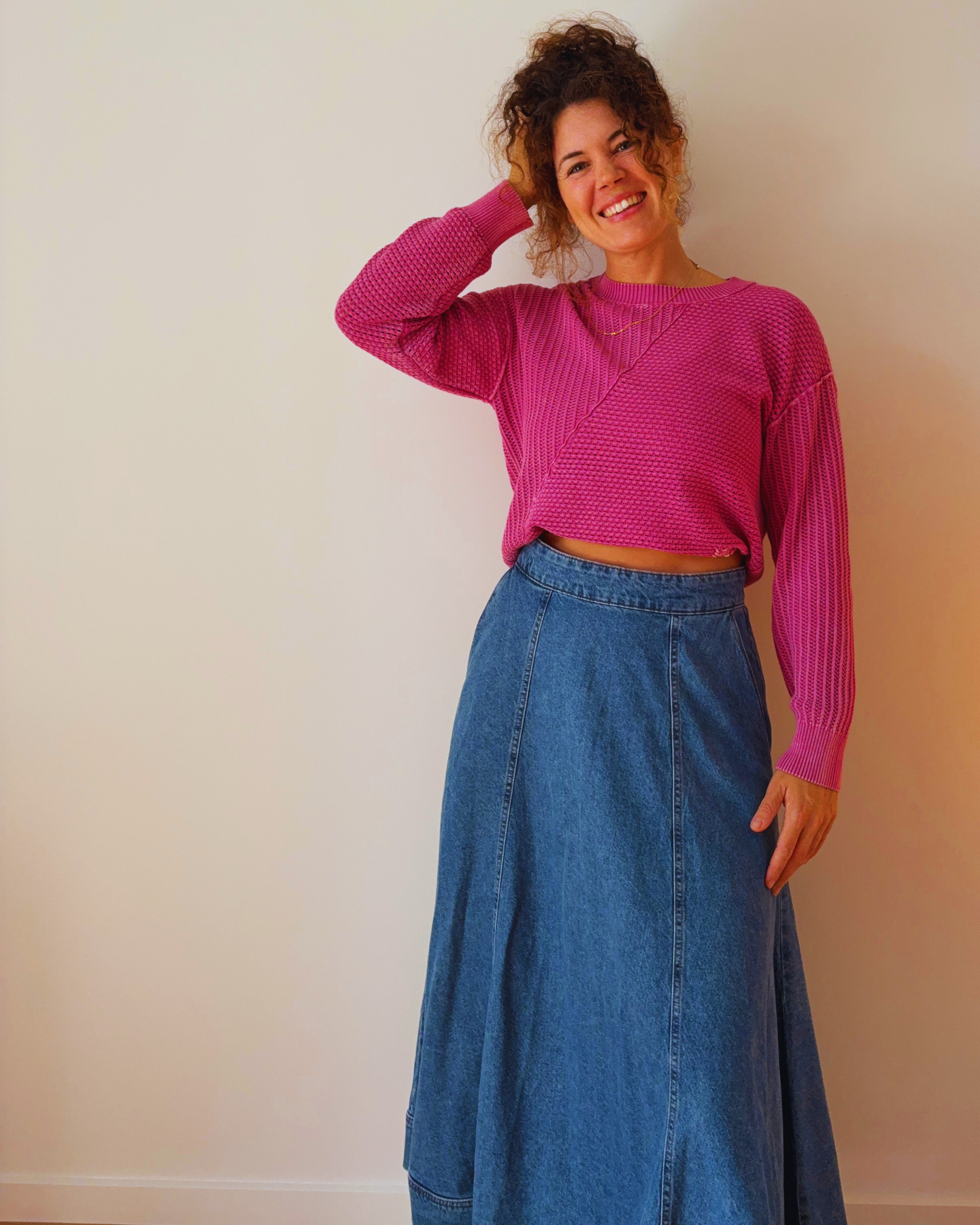 A woman wearing a pink sweater and a long blue denim skirt standing against a plain, light-colored wall, smiling and touching her hair with her right hand.