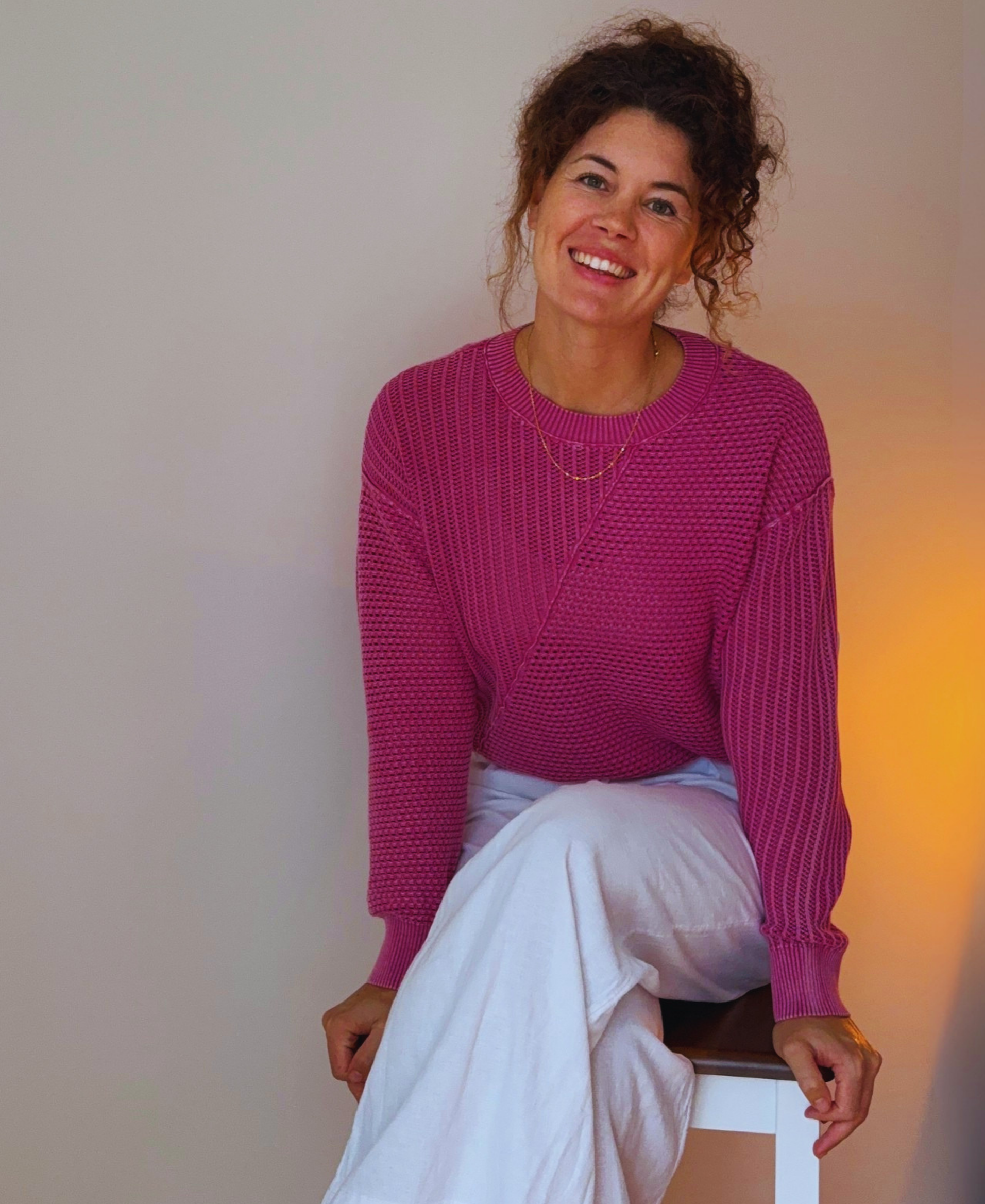 A woman with curly brown hair, wearing a pink knitted sweater and white pants, sitting on a chair, smiling at the camera, against a plain neutral background.