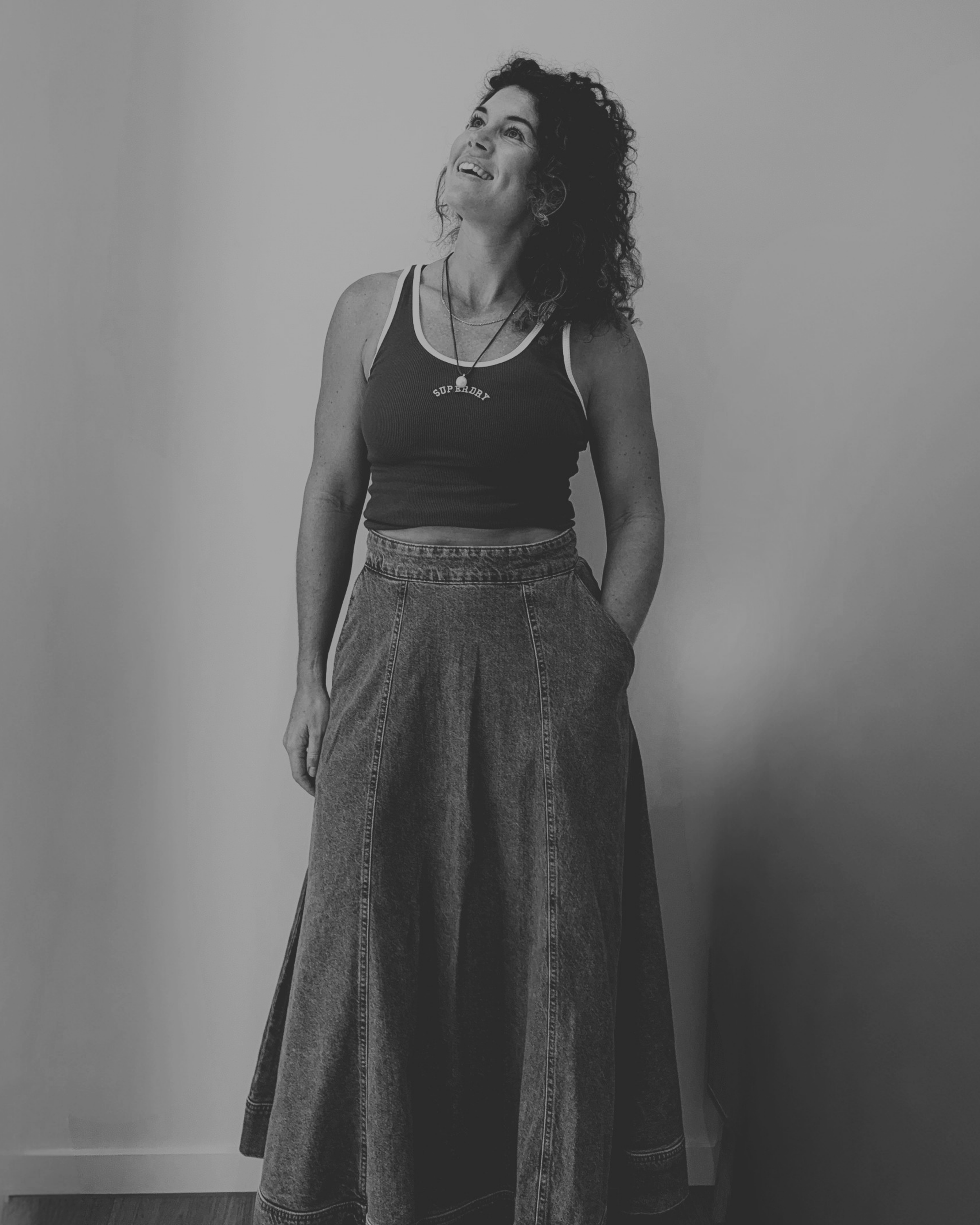 A woman with curly hair wearing a sleeveless top and high-waisted jeans, standing against a plain wall and looking slightly upward with a smile.
