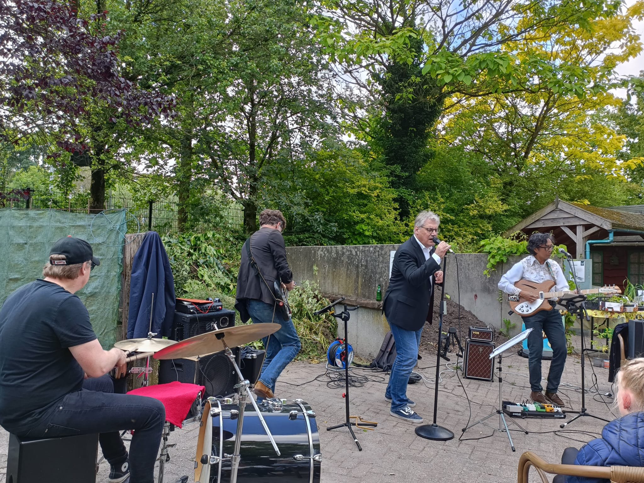Een groep muzikanten voert een optreden uit in een tuin, omringd door bomen met groene bladeren, met een man die piano speelt en een vrouw die gitaar speelt, terwijl anderen opdrummen of luisteren.