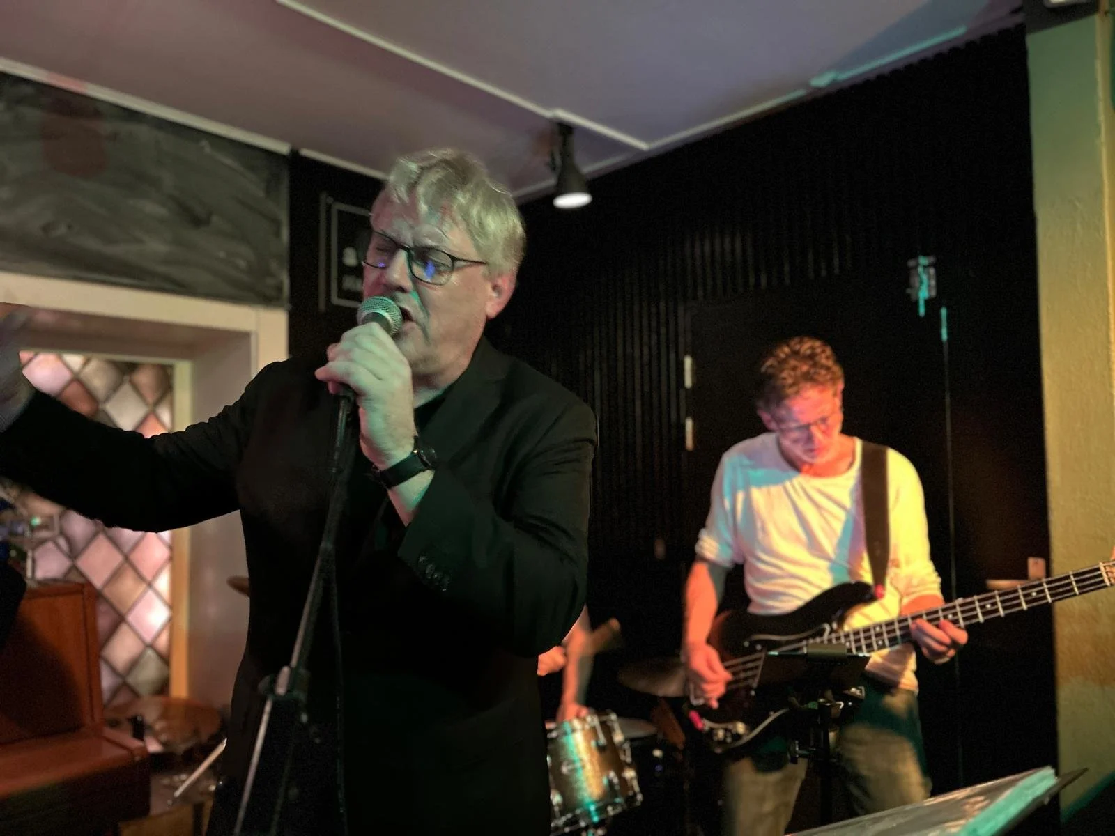 Twee muzikanten spelen in een donkere kamer: een man met blond haar, bril en een zwart pak zingt met een microfoon, terwijl een andere man met kort haar en een lichte trui gitaar speelt.