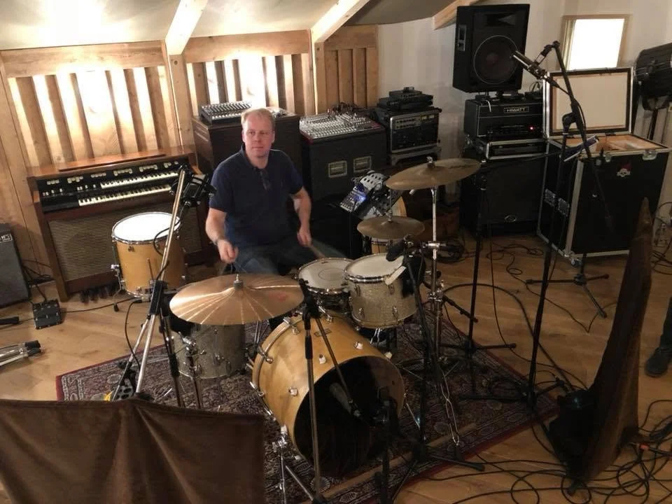 Mannelijke drummer zit naast een drumstel in een geluidsstudio, omringd door audioapparatuur en muziekinstrumenten.
