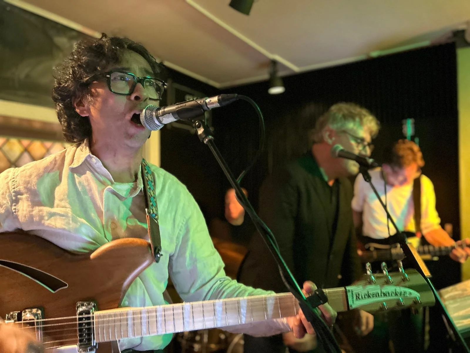 Drie muzikanten spelen live muziek. De man op de voorgrond speelt gitaar en zingt. De andere twee mannen op de achtergrond spelen ukulele en keyboard.