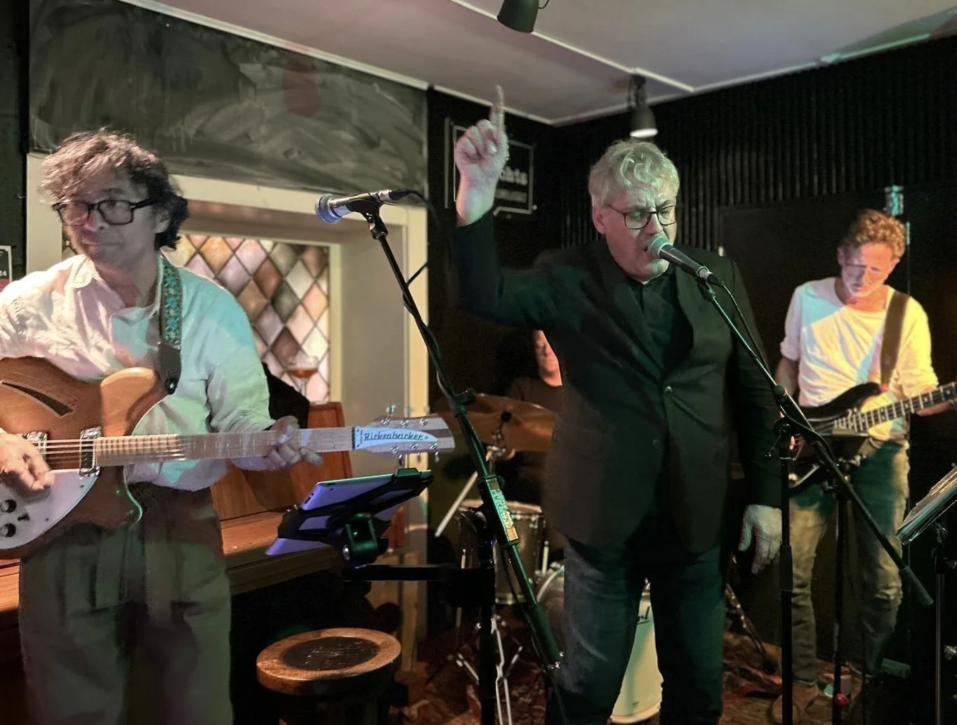 Mannen die muziek maken en zingen in een donkere sfeer, met een gitaar en microfoons, in een kleine podiumsetting.