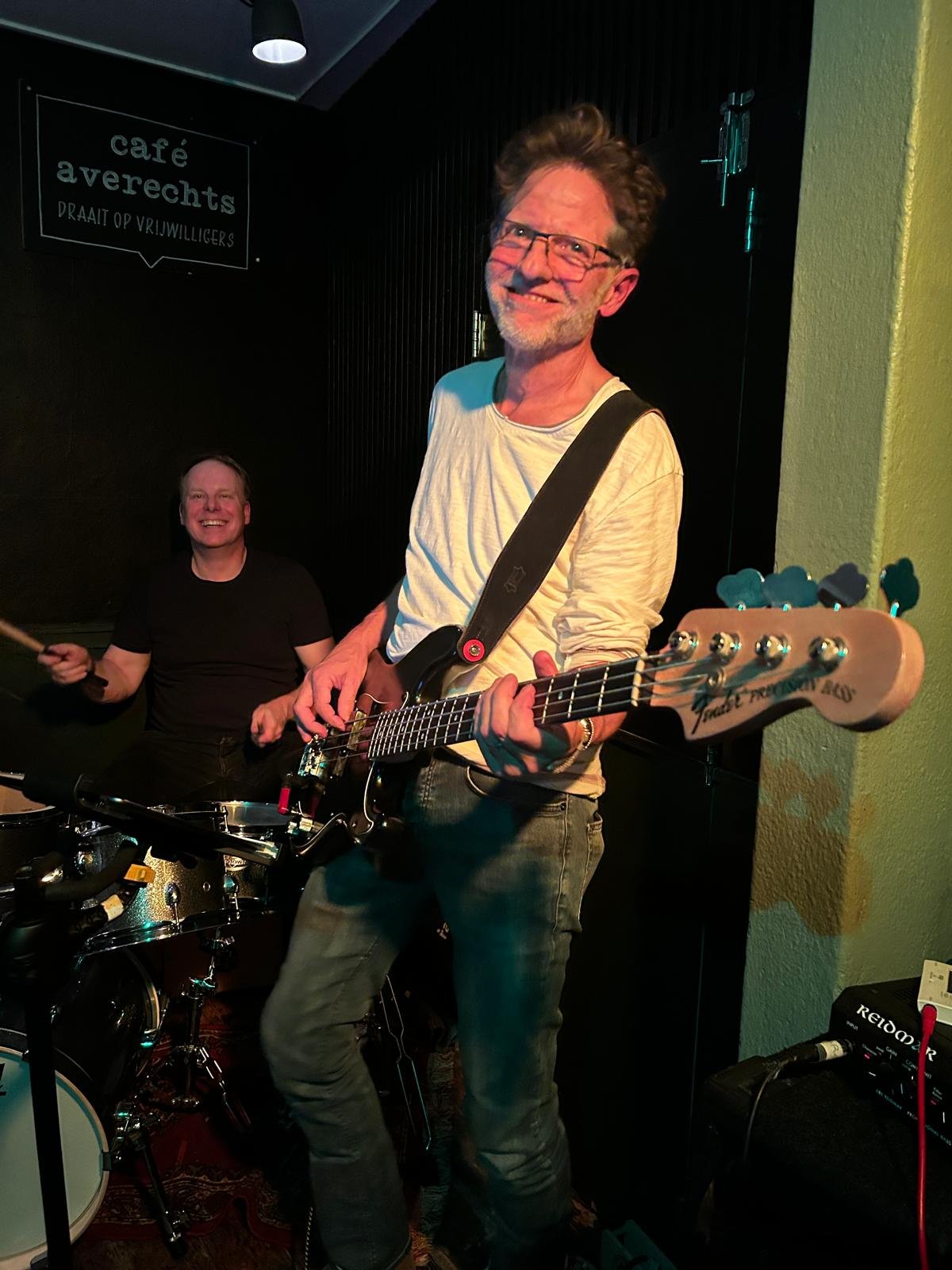 Twee mannen spelen muziek in een donkere ruimte. De man op de voorgrond speelt basgitaar en de man op de achtergrond speelt drums. Er is een bord aan de muur met de tekst 'café asynchronously draait op vrijwilligers'.