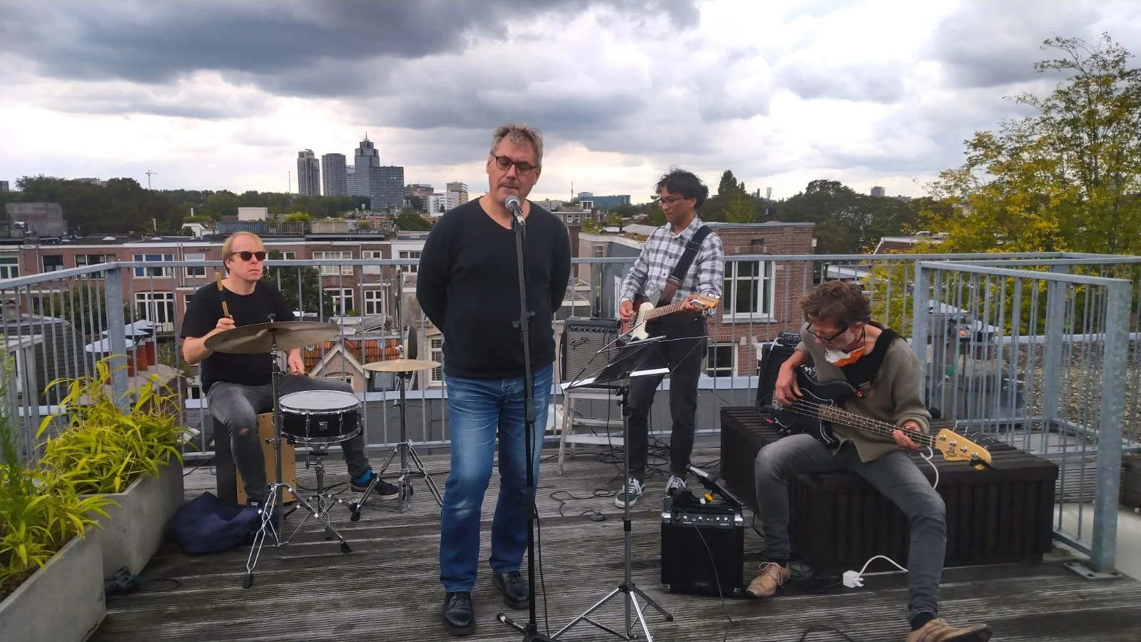 Een band met vier muzikanten op een dakterras speelt muziek, onder een bewolkte hemel. De groep bestaat uit een man met een microfoon, een drummer, een gitarist en een bassist.