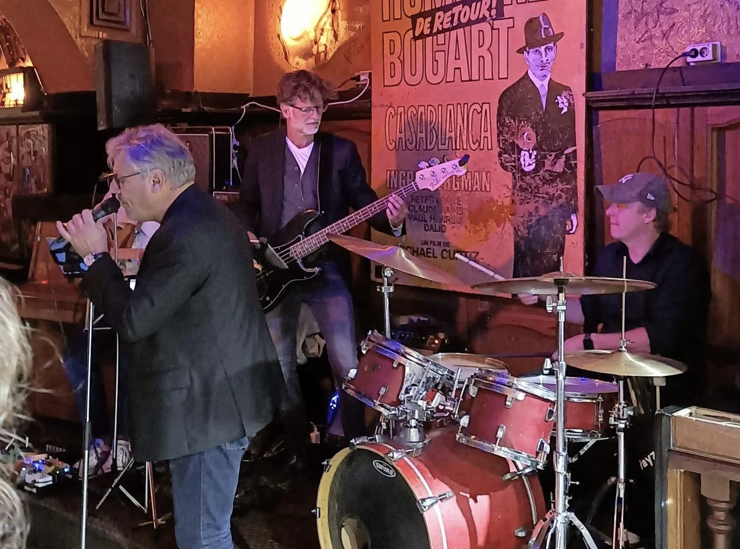 Een muzikale band speelt live in een café of bar. Er zijn drie muzikanten: een zanger met een microfoon, een bassist en een drummer. Op de achtergrond hangt een poster met de tekst 'Hommage de retour! BUDGART CASABLANCA'.