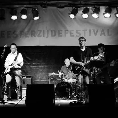 Een band speelt live op een podium tijdens een festival, met vier muzikanten, twee gitaristen, een drummer en een bassist, onder podiumverlichting.