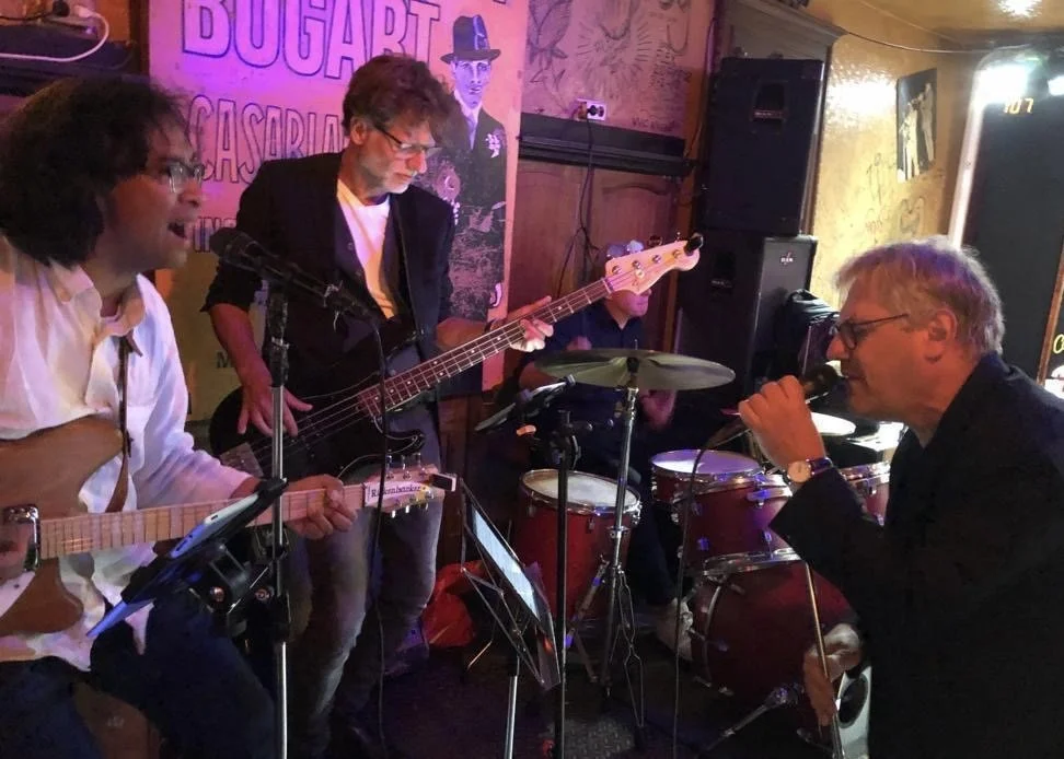 Een live muziekoptreden met vier muzikanten in een gezellige bar of cafe. Twee gitaristen, een bassist, en een zanger die ook drums bespeelt, onder kleurrijke verlichting.