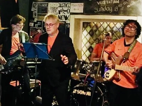 Drie mensen spelen muziek in een band, met een instrumenten zoals gitaar en drums, in een café of kleine muziekruimte.