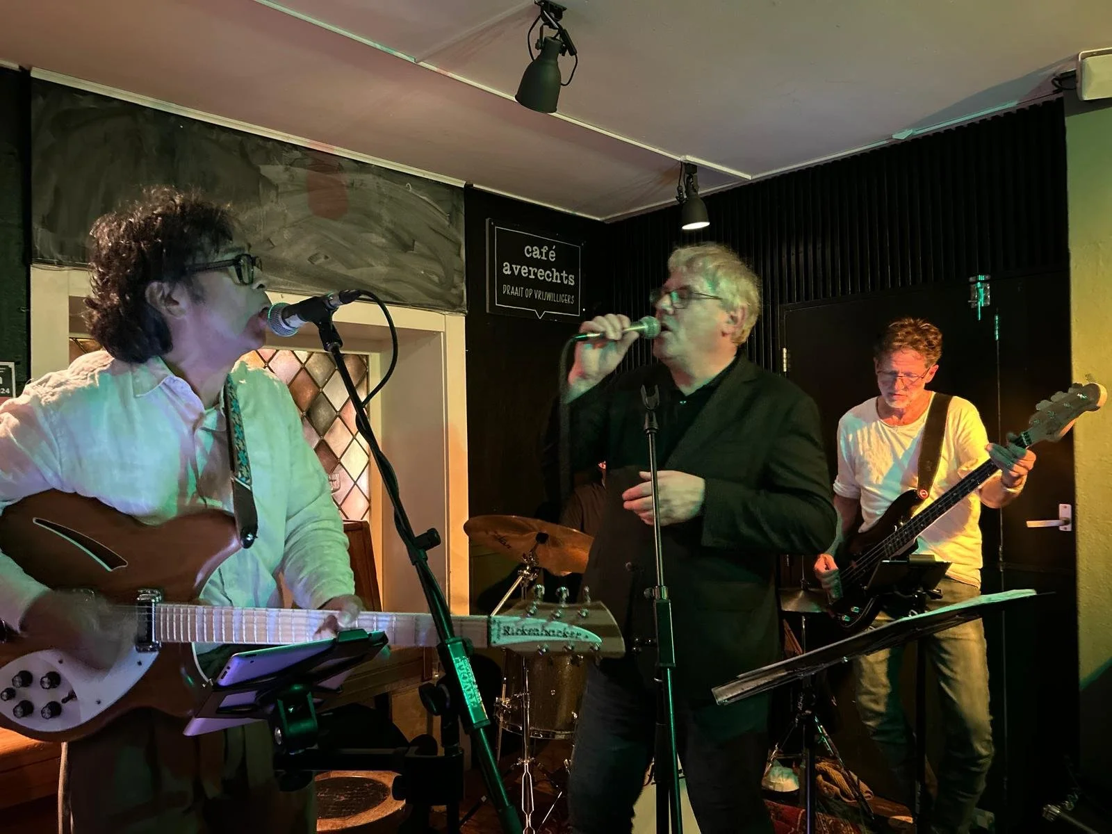 Drie muzikanten spelen live in een café, één met een gitaar, één met een microfoon en een ander op basgitaar, in een gezellige muzieksetting.
