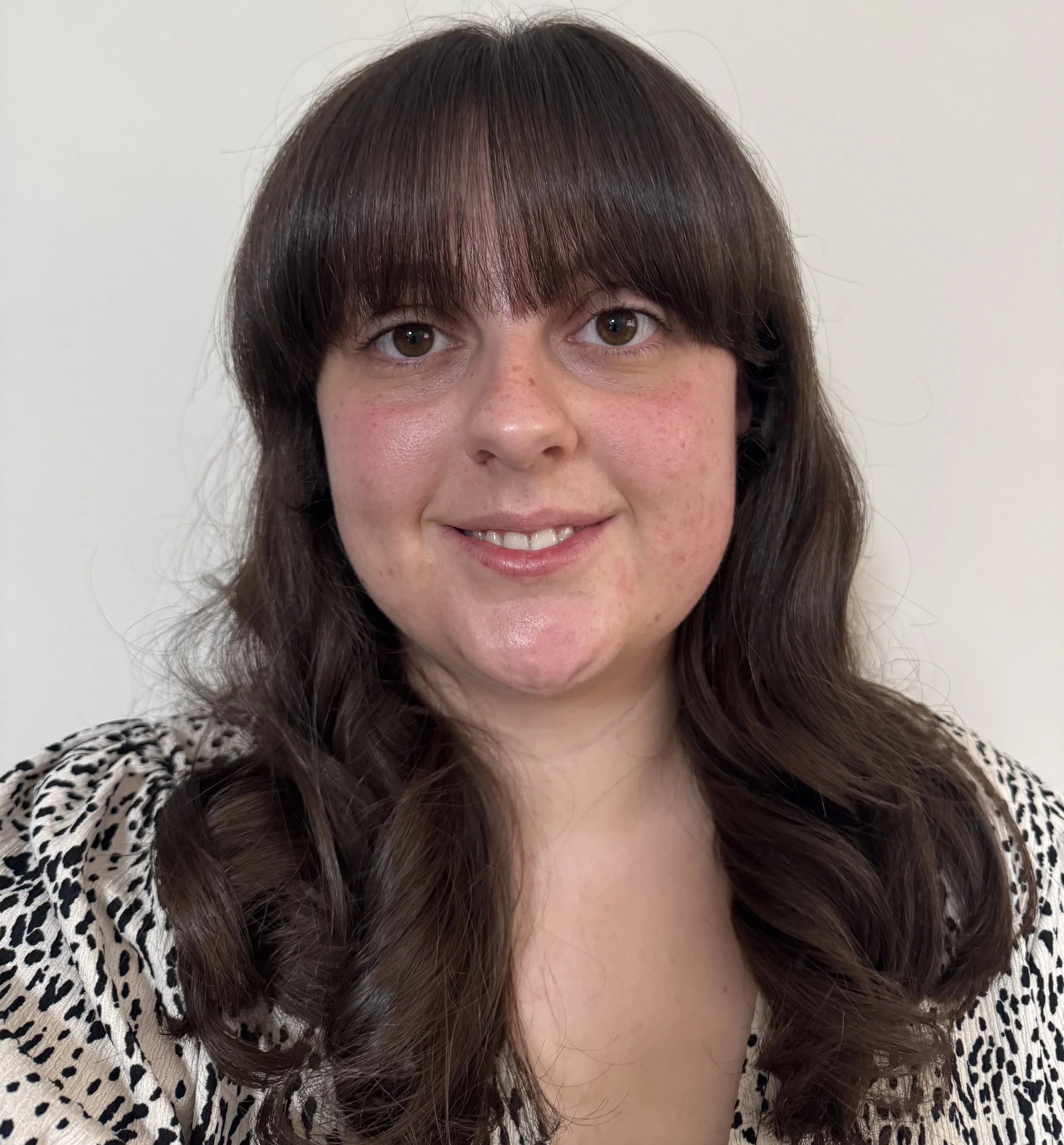 A woman with brown hair and brown eyes wearing a leopard print top, smiling slightly in front of a plain white background.