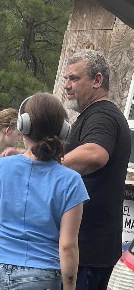 A man with gray hair and a beard talking to a woman wearing large headphones, with a girl in the background, outdoors near a wooden structure and trees.