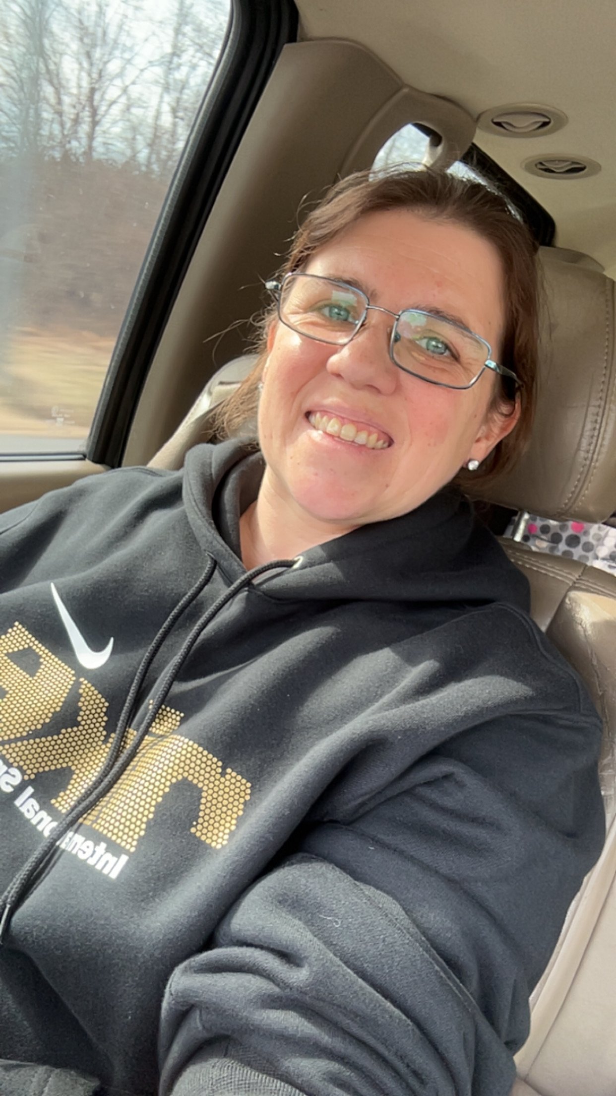A woman with glasses smiling while sitting in the backseat of a car.