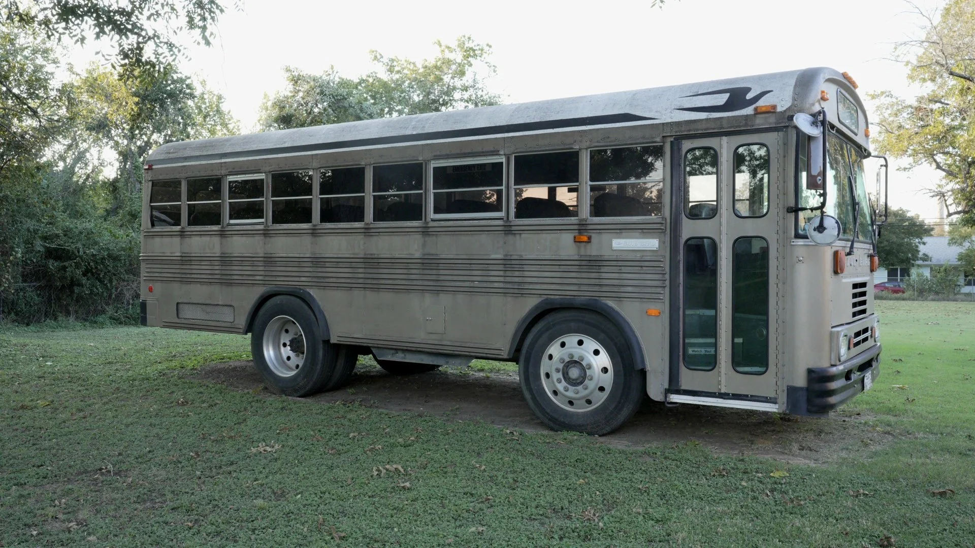 An old, silver bus parked on a grassy area with trees and a house in the background.