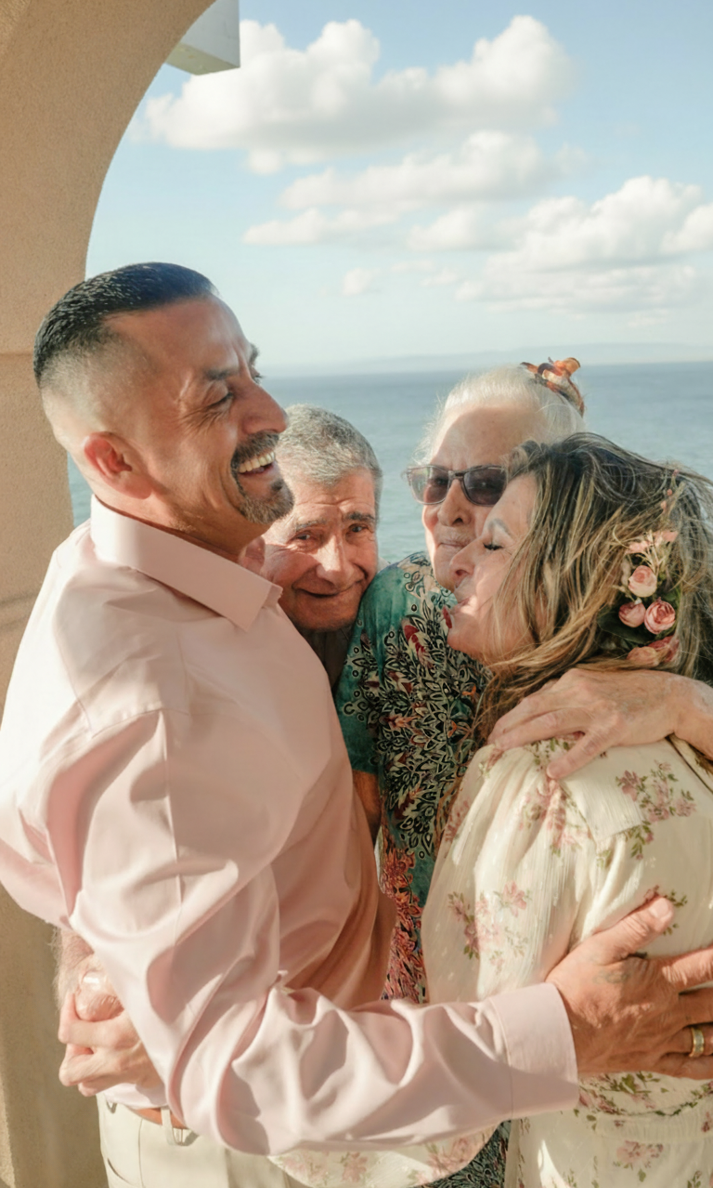 A man and woman are hugging and smiling outdoors by the water with a blue sky and clouds in the background. The man has dark hair and a beard, wearing a pink shirt. The woman has long hair with flowers, wearing glasses and a floral dress.