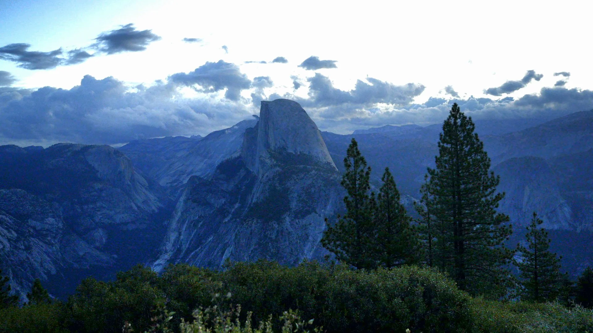 Mountain landscape with large rock formation, pine trees in foreground, cloudy sky.