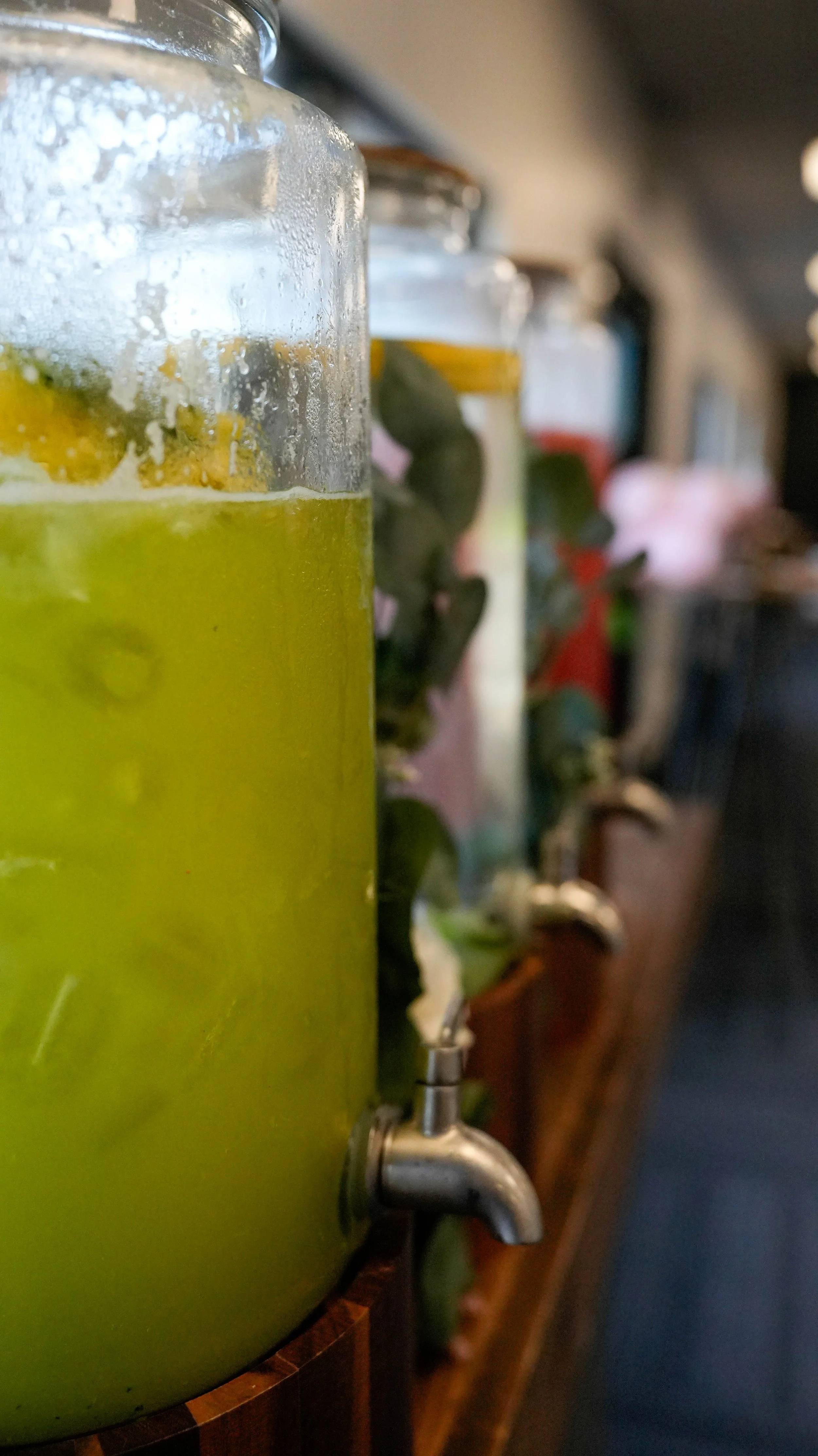 Close-up of a large glass jar filled with a green liquid, placed on a wooden surface with a metal spigot at the bottom. Other similar jars are blurred in the background.