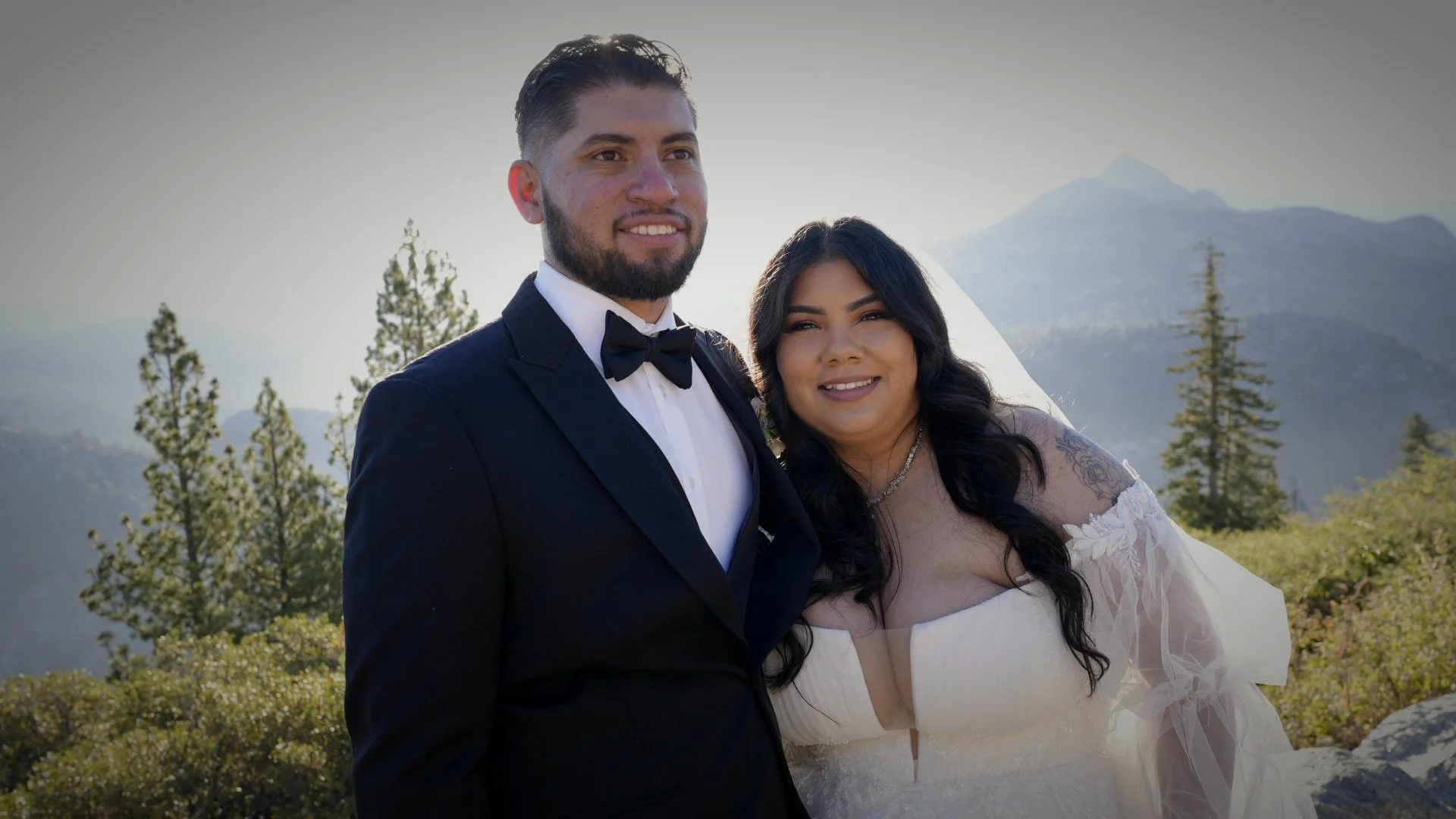 A bride and groom standing outdoors in a mountainous area during daytime, sunlight behind them, near trees and green scenery, both smiling.