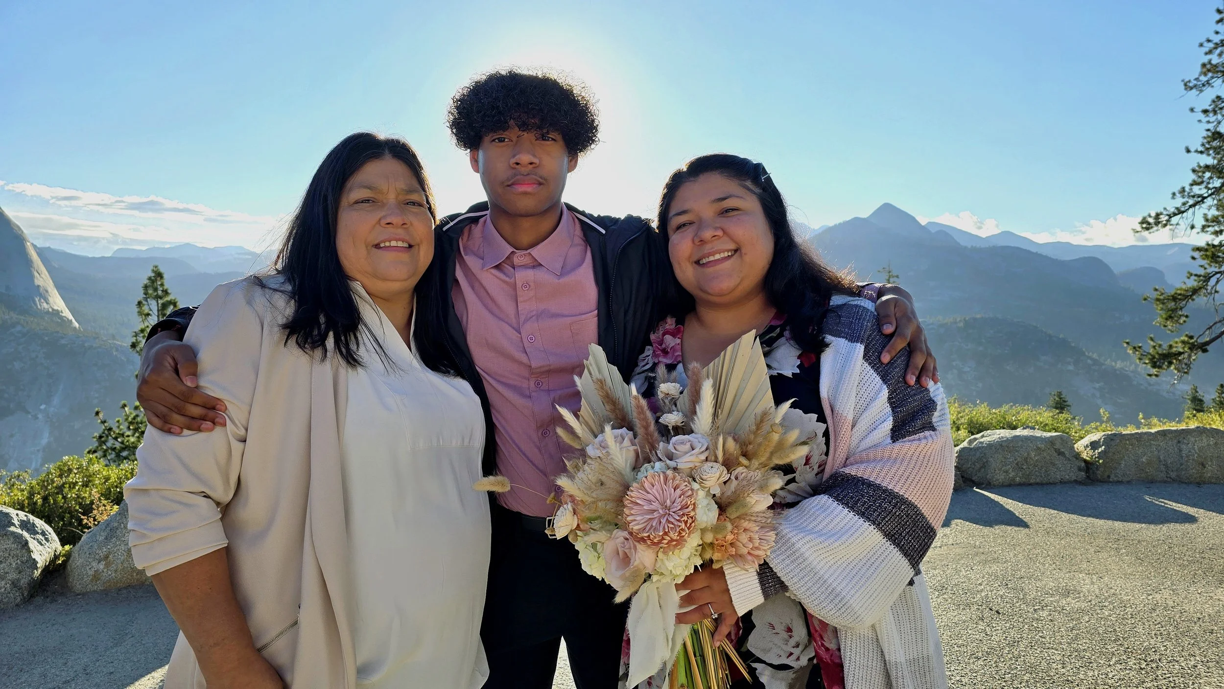Three people standing outdoors with mountains in the background, embracing and smiling, with a woman holding a bouquet of flowers.